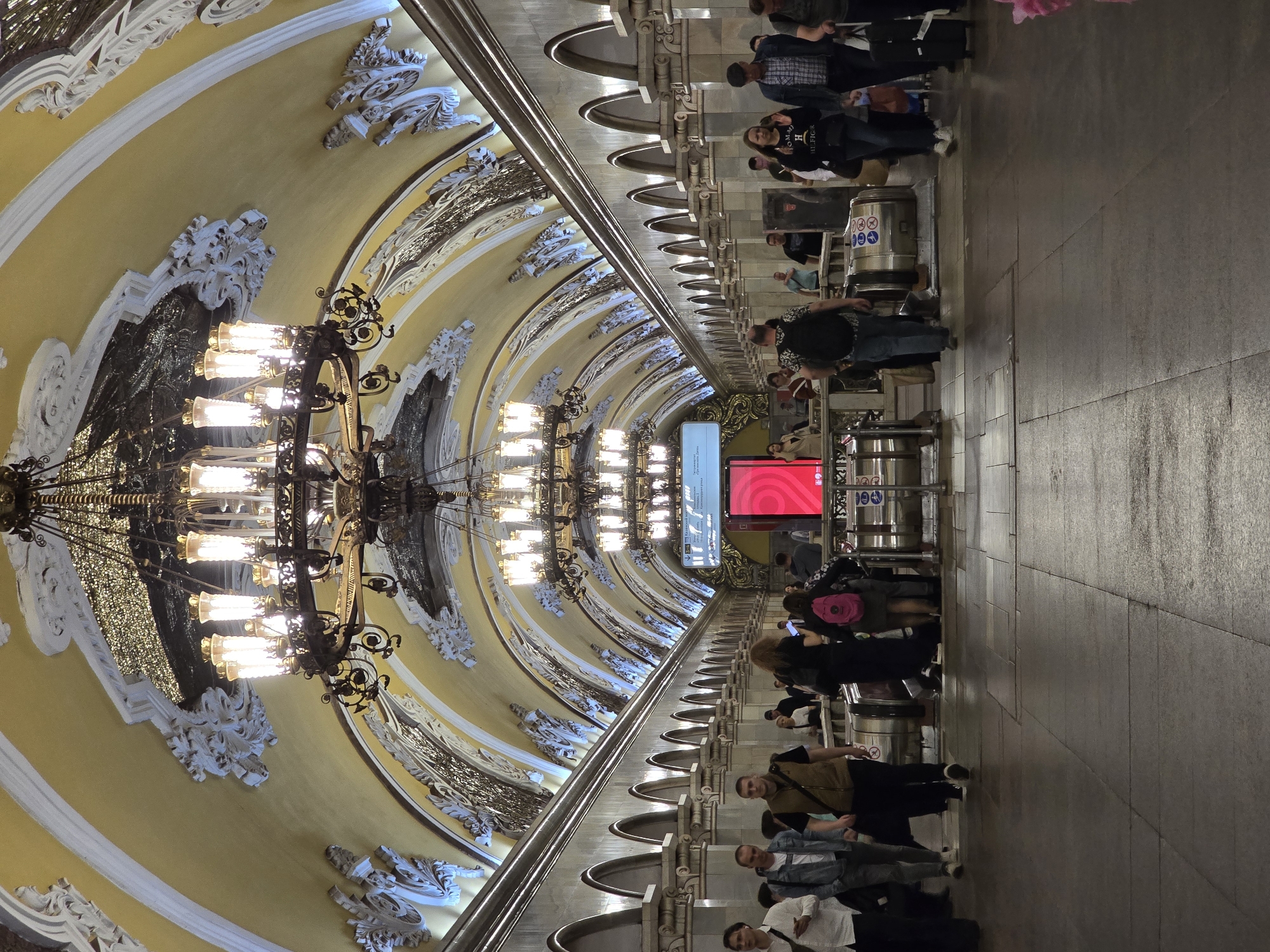 Captured indoors, the image depicts a busy subway station in Moscow, Russia. The station is characterized by an elaborate, vaulted ceiling, ornamented with intricate stucco details and illuminated by ornate chandeliers. People, standing and walking, crowd the tiled platform area, moving past the turnstiles. A digital sign, displaying both information and advertisements, is visible in the distance. The scene provides a glimpse into Moscow's public transportation system, emphasizing its grandeur and daily activity.