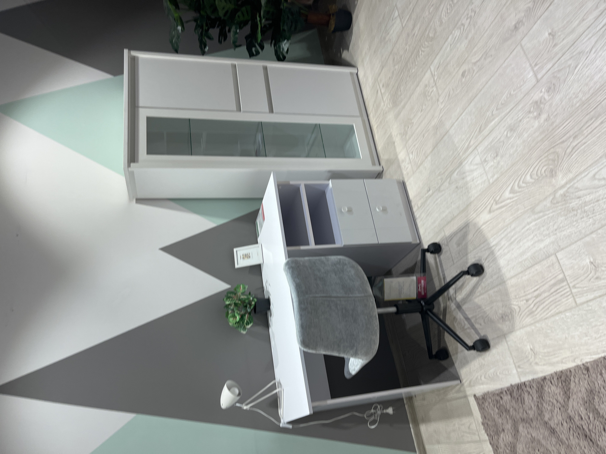 The image shows a modern home office setup. A white desk with a minimalist white lamp and a potted plant is positioned against a wall decorated with a geometric pattern of grey, white, and mint green triangles. A grey office chair with a wheeled base sits at the desk. To the right of the desk is a tall white cabinet with glass-fronted doors, displaying shelves inside. Next to the cabinet, a large potted plant with dark green leaves is visible. The floor is covered with light-colored laminate flooring, and a corner of a brown rug is visible in the lower left of the frame. The overall impression is one of a clean, organized, and contemporary workspace. No specific landmarks or elements in the image definitively place the scene in Kharkiv, Ukraine.