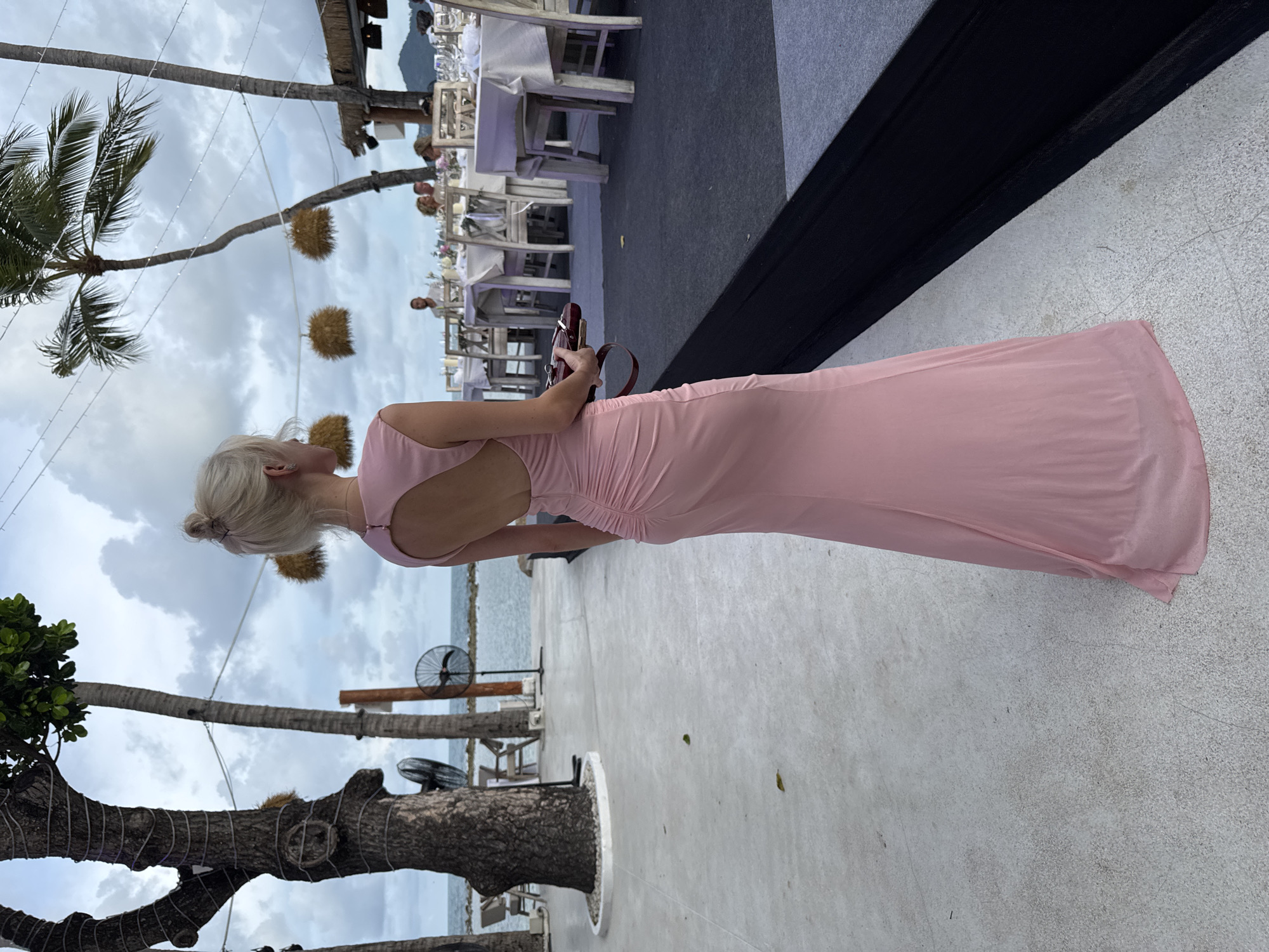A woman in a pink dress is standing on a light-colored surface, possibly concrete, in บ้านบางรักษ์, Thailand. She is facing away from the camera and holding a small, dark-colored object, possibly a clutch or phone case. In the background, there are palm trees, dining tables set for a meal, and what appears to be a beach or coastal area. The sky is overcast.