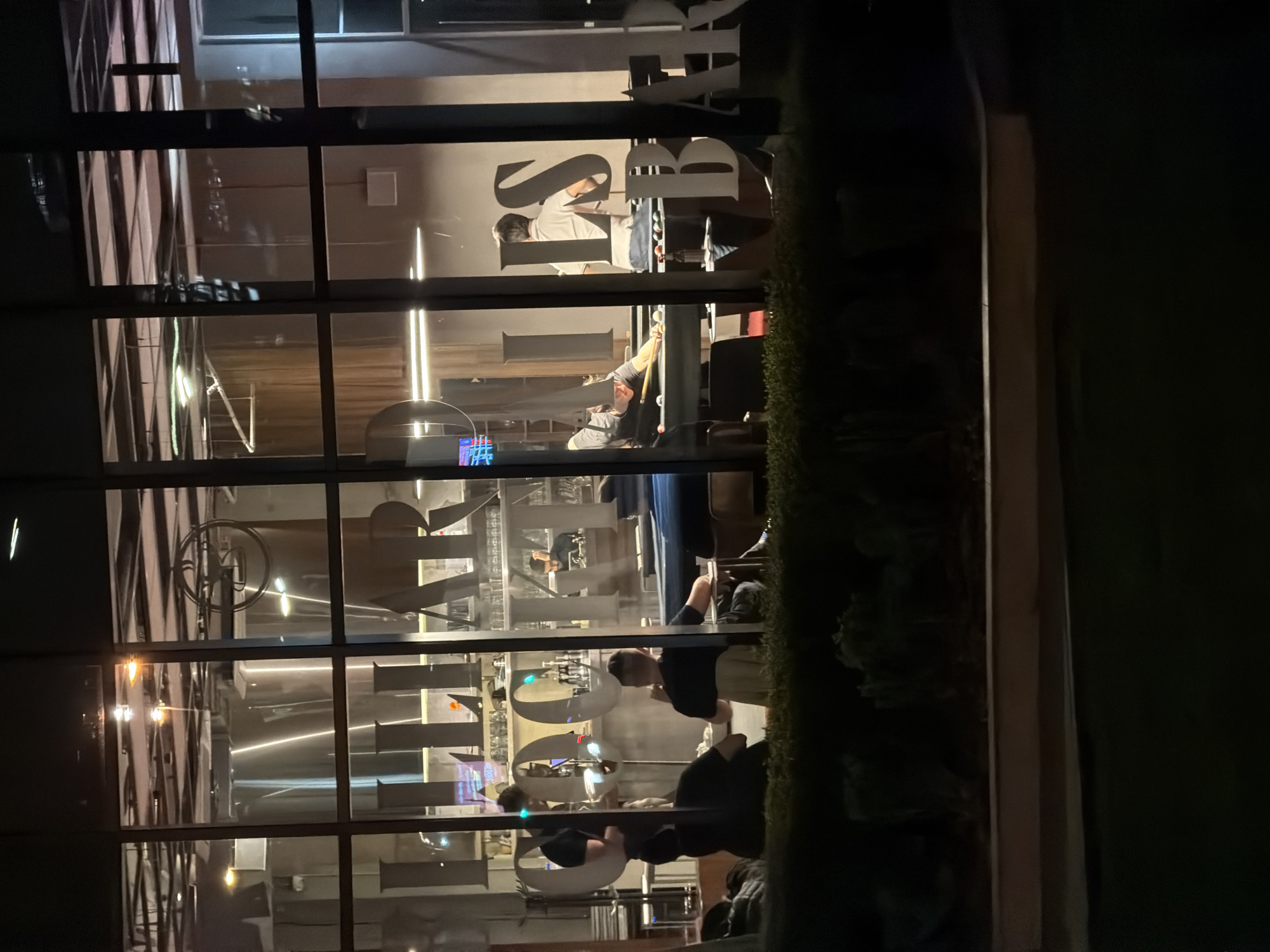 The image displays the interior of a billiard and cocktail bar viewed from outside at night through large glass windows. The establishment is the Kabinet Brewery & Billiard Cocktail Bar, located at Autoput za Novi Sad 150, Zemun, Belgrade, Serbia.
Inside, bright artificial lighting illuminates the scene. Prominent white lettering affixed to an interior wall spells out "BILLIARD COCKTAILS BAR." In the background, large, gleaming stainless steel industrial tanks, indicative of a brewery, are visible. Approximately 6-8 individuals are present.
To the left, a man in a dark shirt stands behind a bar counter, holding a white plate or bowl with both hands. Two other men are seated at a table in the immediate vicinity. In the center and right, two men are actively engaged in playing billiards on a table covered with blue felt. One man, wearing a light-colored shirt, is bent over the table, holding a cue stick, preparing to strike the balls. Another man, in a dark shirt, stands near this billiard table. A bottle is visible on a small table adjacent to the billiard players. Further to the right, a portion of a second billiard table is visible. In the deep background, behind another bar section, a man in a dark shirt is also visible.
The exterior foreground consists of a low, dark green hedge or shrubbery, situated above a light-colored, horizontal ledge or wall.