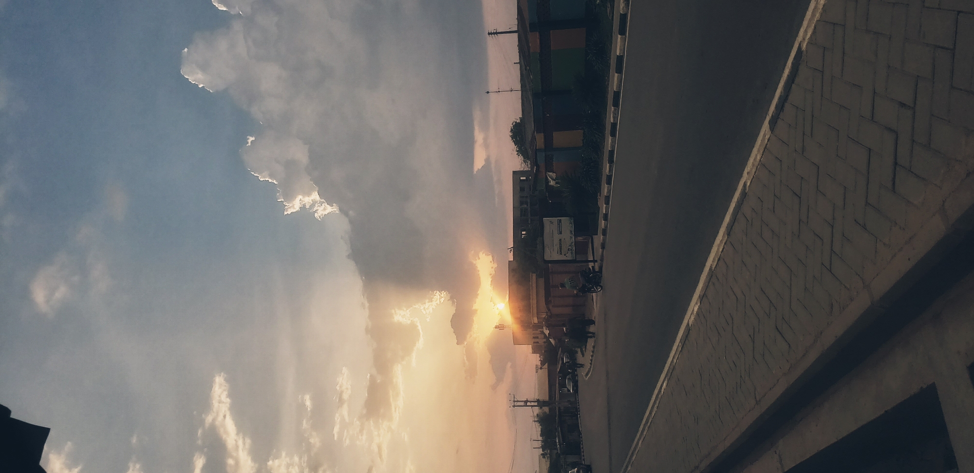 The image, oriented in landscape, captures a vibrant sunset or sunrise over a street in Narayi, Nigeria. The sun, low on the horizon, is the focal point, radiating a bright golden light that breaks through and illuminates scattered clouds, creating a dramatic sky of soft blues, oranges, and yellows.
In the mid-ground, an asphalt road with a distinct white and black striped shoulder or curb stretches from left to right. Along the roadside, various buildings are visible, some with colored panels and others seemingly under construction, nestled amidst sparse vegetation. A motorcycle is parked near a white signboard on the left side of the road, on which the word "ADMISSION" is clearly legible. Other indistinct vehicles and figures are visible further down the street, suggesting daily activity.
The foreground on the right features a prominent concrete wall or barrier with a decorative herringbone brick pattern, indicating an elevated perspective from which the photograph was taken. Utility poles and wires are also faintly visible against the sky. The overall scene evokes the serene yet active ambiance of an urban fringe at the transition between day and night.