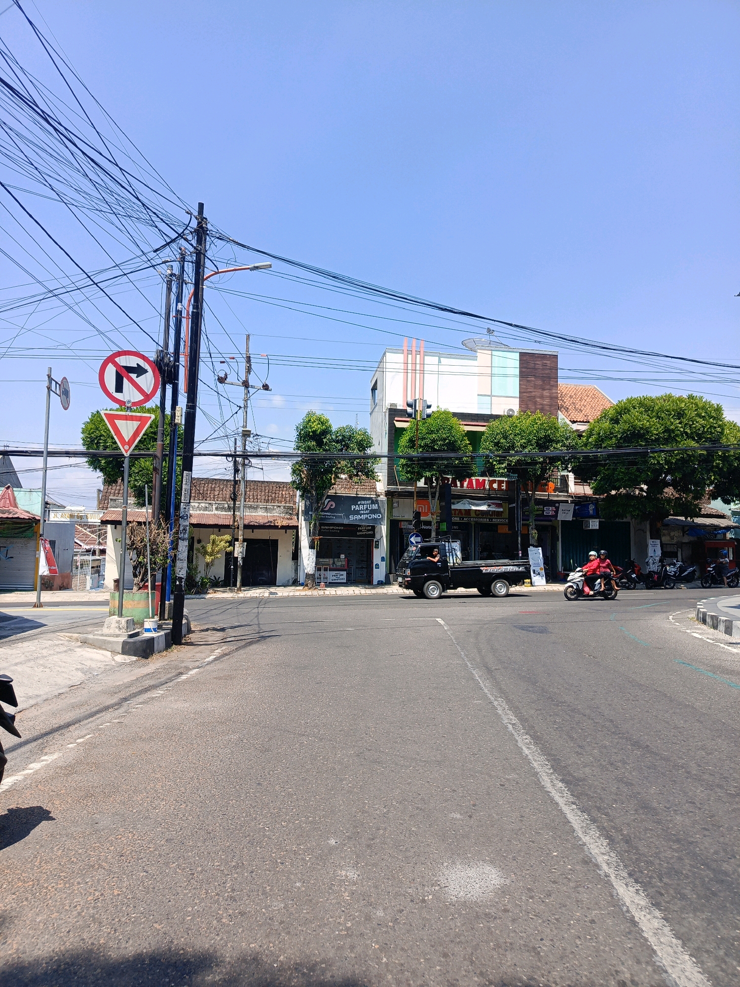 The image shows a street intersection in Magetan, Indonesia. The sky is clear blue. On the left side of the road, there is a traffic sign indicating "no right turn". There is a traffic light pole with many wires connected to it, as well as a street lamp. In the background, there are several buildings, some with awnings and shops. One building has a sign that says "PARFUM SAMPOERNA". A black pickup truck is driving through the intersection. To the right, two people are riding a red motorcycle. In the distance, other motorcycles are parked. The road is asphalt and marked with white lines.