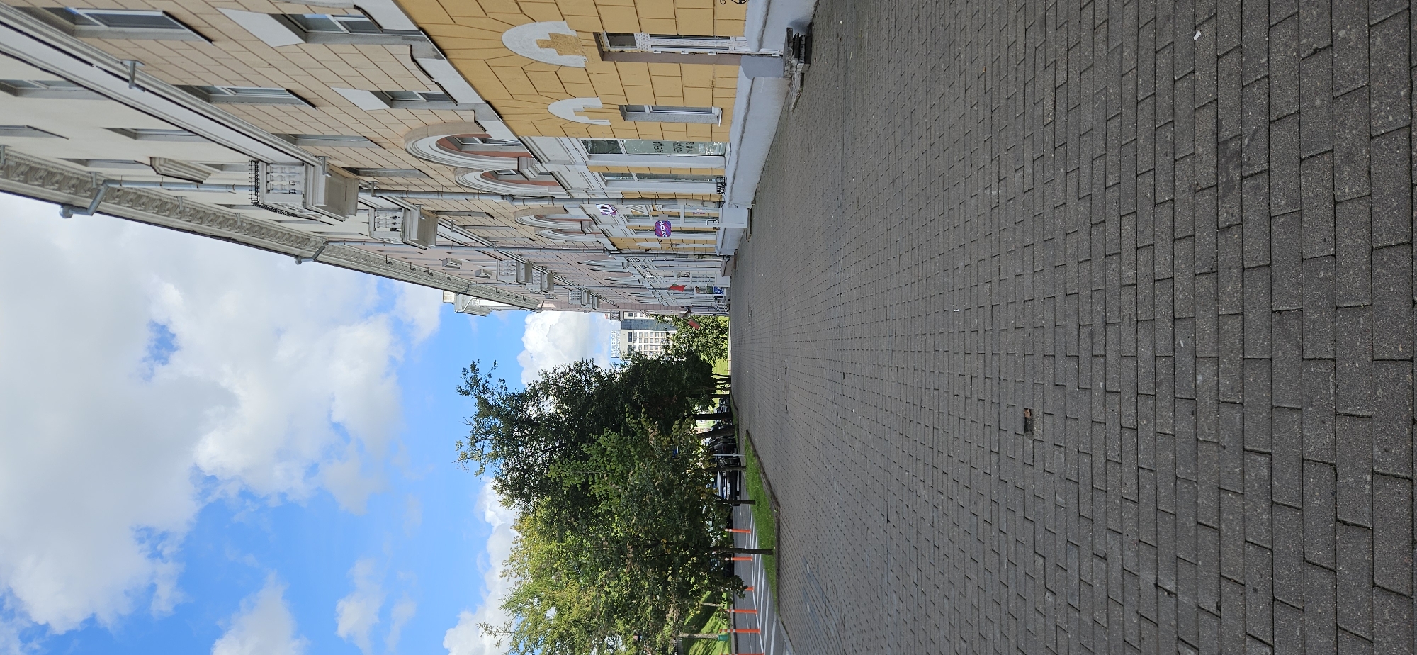 The image shows a view down a pedestrian sidewalk paved with grey rectangular bricks, bordered by a street with parked cars on the left and a long, yellow building on the right. Trees line the left side of the sidewalk, separating it from the street. The building on the right is multi-story with a decorative facade, featuring arched windows and balconies. The sky above is blue with scattered white clouds. In the distance, more buildings are visible. A sign for "OZON" is visible on the ground floor of the yellow building, indicating a commercial area. The scene appears to be on a clear, daytime, likely summer or early autumn, in an urban environment. This location is identified as Victory Square in Vitebsk, Belarus, a prominent historical and public space.