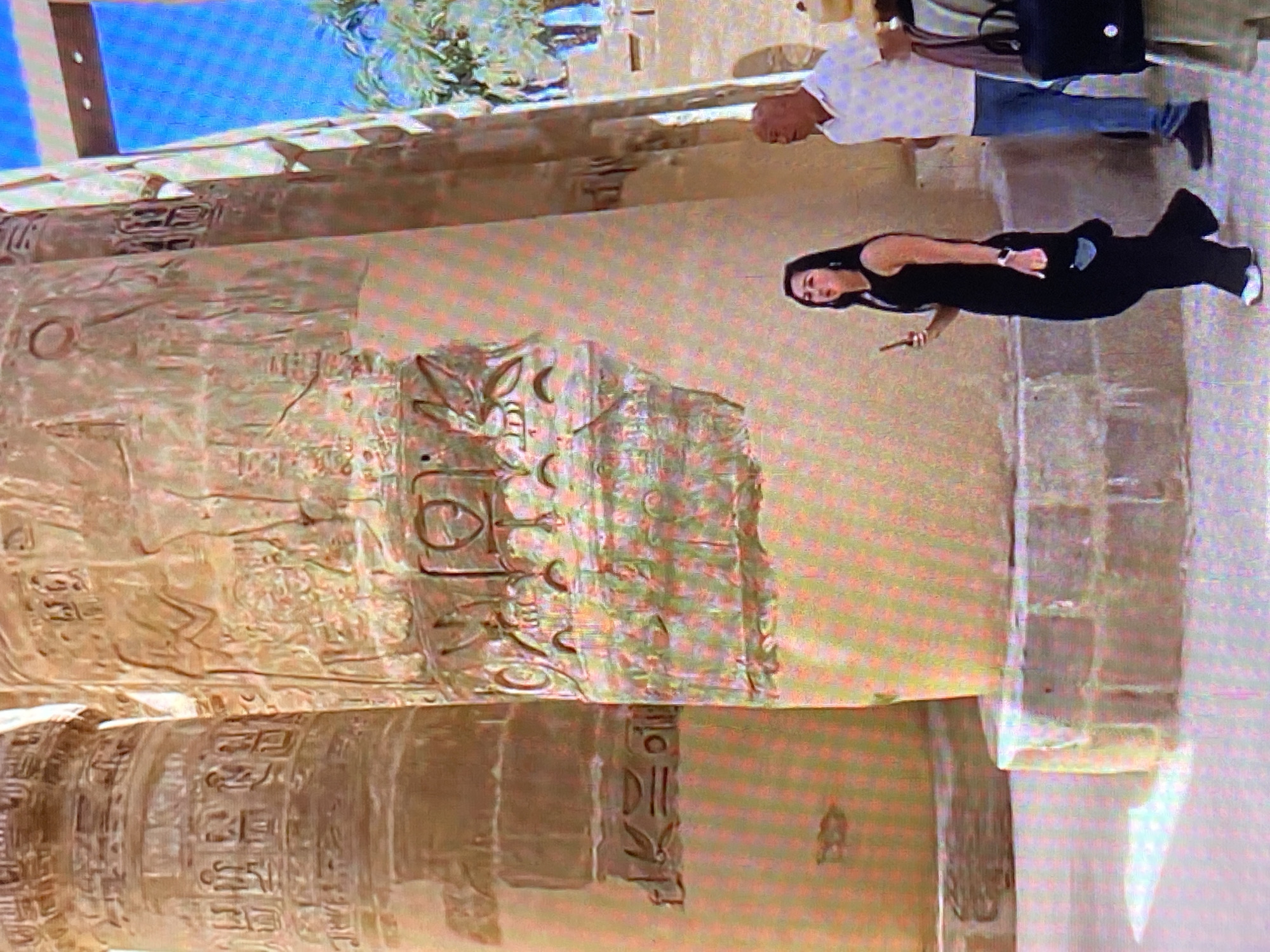 The image shows a scene at the Karnak Temple in Luxor, Egypt. A large, intricately carved column dominates the left side of the frame, displaying Egyptian hieroglyphs and relief carvings. To the right, a woman dressed in black points with her right hand at the column while holding a small black object in her left. An older man in a white shirt and blue jeans stands behind her, observing the column. A black bag hangs from his right shoulder. The scene is outdoors, with bright light and foliage visible in the top of the frame. The ground appears to be a light-colored stone or concrete.