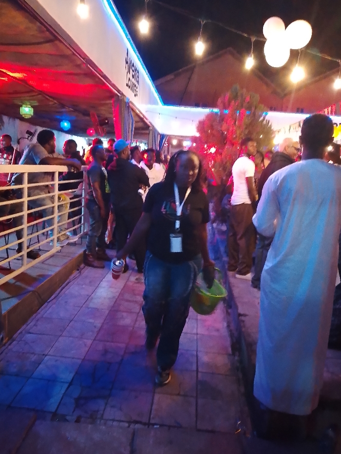 The image displays a nighttime social gathering within an outdoor or semi-outdoor venue identified by a visible sign as "Arena Lounge" in Narayi, Nigeria. The area is lit by overhead string lights, blue LED strip lighting along an awning, and general red ambient illumination. Approximately fifteen to twenty individuals are present. In the immediate foreground, a dark-skinned woman walks forward, carrying a silver can with red details in her right hand and a green bucket in her left. She wears a dark shirt with a graphic, dark jeans, and an ID badge suspended from a lanyard around her neck. To her right, a person in a light-colored traditional garment is partially visible from behind. On the left side, several men are gathered near a white railing, with one male leaning on it. In the mid-ground, other groups of individuals stand and converse. A small, illuminated tree with reddish foliage is centrally located on the right side. The ground is paved with rectangular tiles. The scene indicates a leisure or event setting.