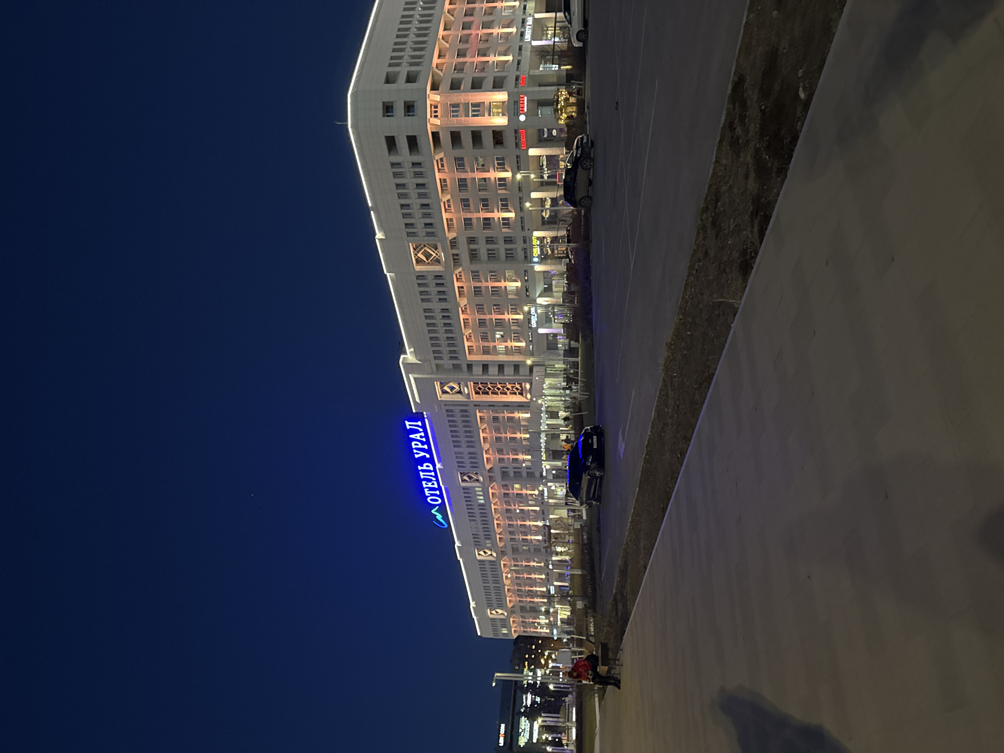 Here's a forensic description of the image:
The image shows a street scene at night in Perm, Russia. The Hotel Ural is prominently visible with its blue neon sign in Russian Cyrillic script. The building has a rectangular form, multiple floors, and regular window arrangements with ornamental dividers. The street includes a broad paved area or sidewalk in the right-hand side of the picture. A narrow landscaped strip lies between the road and the pavement. Lighting from street lamps and the building illuminates the scene. A person is visible on the edge of the pavement. A dark vehicle is parked close to the hotel. The sky is dark blue.