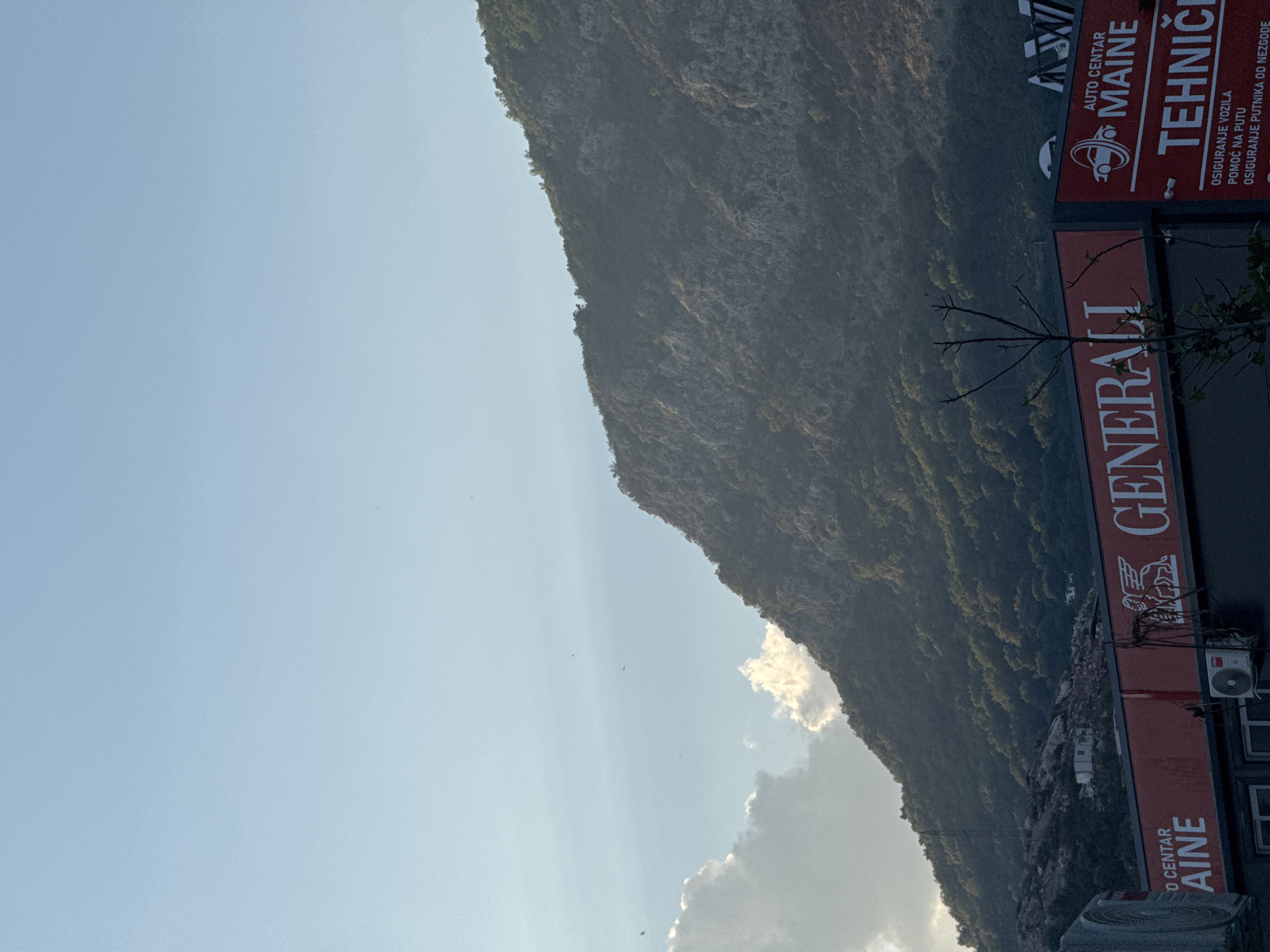The image captures a portion of the Budva coastline in Montenegro, featuring a rugged, tree-covered mountain slope ascending into a pale blue sky with scattered clouds. In the foreground, a red awning with white lettering displays "GENERALI," indicating a business. To the right, another business sign, also red with white text, reads "AUTO CENTAR MAINE" and below it "TEHNIČKI," with further text detailing services such as "OSIGURANJE VOZILA" (vehicle insurance). Two white air conditioning units are visible mounted on the building. The scene is illuminated by the soft light of either dawn or dusk, casting a gentle glow on the landscape and buildings. No people are present in the frame.