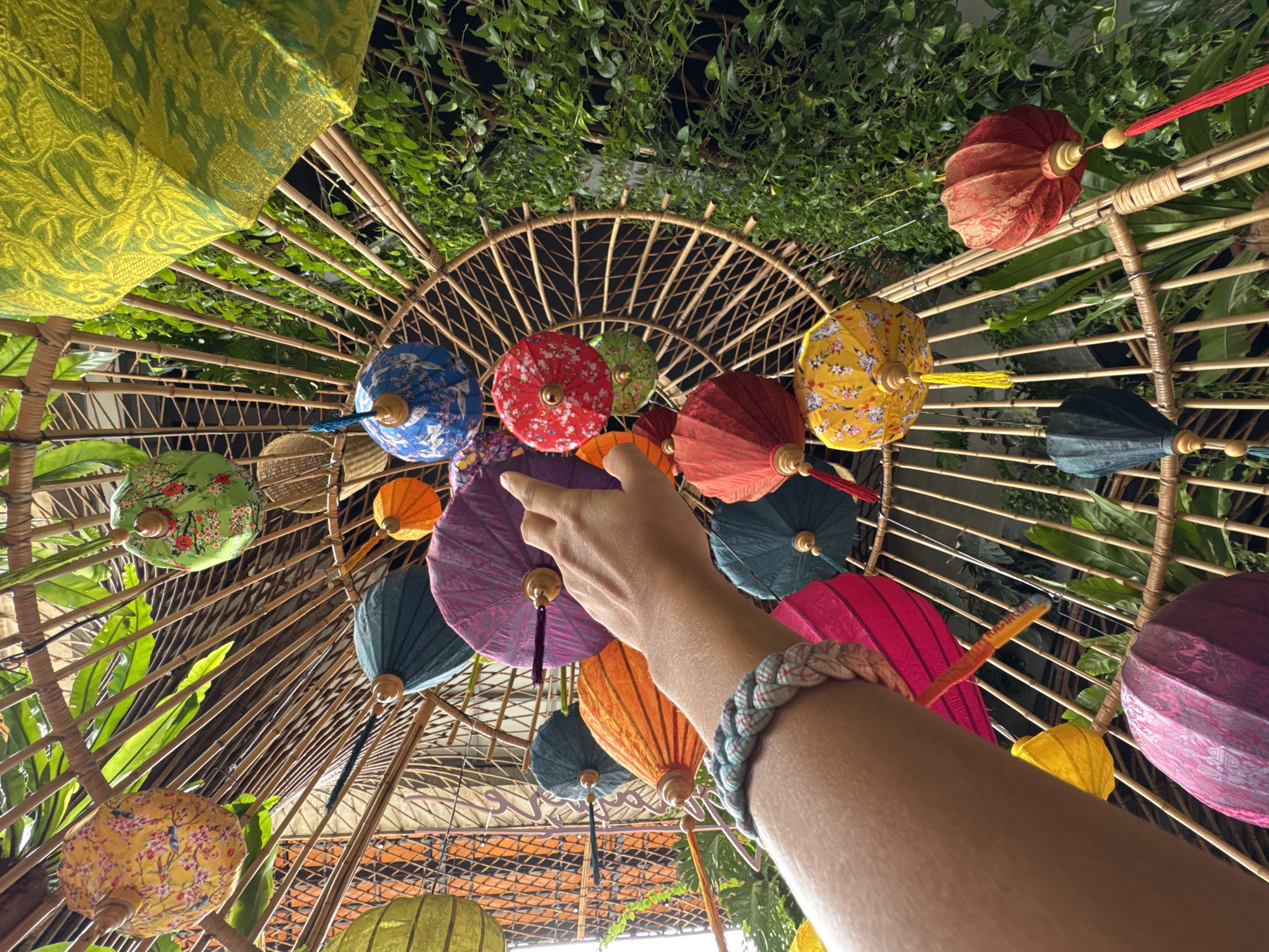 The image captures a low-angle view of a collection of decorative, colorful Vietnamese lanterns suspended within a bamboo structure. A person's arm, adorned with a braided bracelet, extends into the frame, with a finger pointing towards a purple lantern. The lanterns are varied in color, including yellow, green, blue, red, orange, and pink, and many are decorated with intricate patterns and tassels. Lush green foliage surrounds the bamboo structure, suggesting an outdoor or semi-outdoor setting. The scene appears to be in Nha Trang, Vietnam, as indicated by the presence of traditional Vietnamese lanterns. The overall impression is one of vibrant cultural decoration.