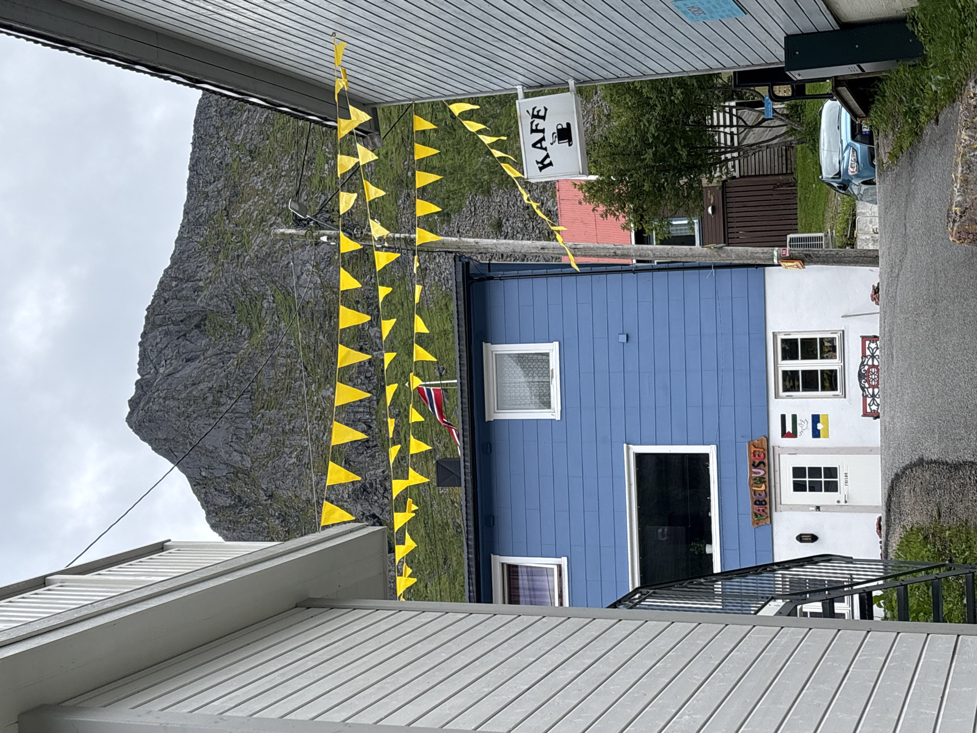 In Gryllefjorden, Norway, a street scene unfolds with a blue building prominently featured, adorned with yellow pennant banners strung overhead and a "KAFÉ" sign. The building has white trim around its windows and a white lower story, on which the sign "FABELHUSET" is displayed above a white door. To the right of the door, there are three flags: a Norwegian flag, a Palestinian flag, and a Ukrainian flag. A steep, rocky mountain looms behind the building, with patches of green grass. On the left, a gray-sided building with white trim partially obscures the view. A black metal staircase descends along the side of the gray building. A light blue car is parked on the street, visible to the right of the blue building, with a mountain and trees in the background. The sky is overcast.