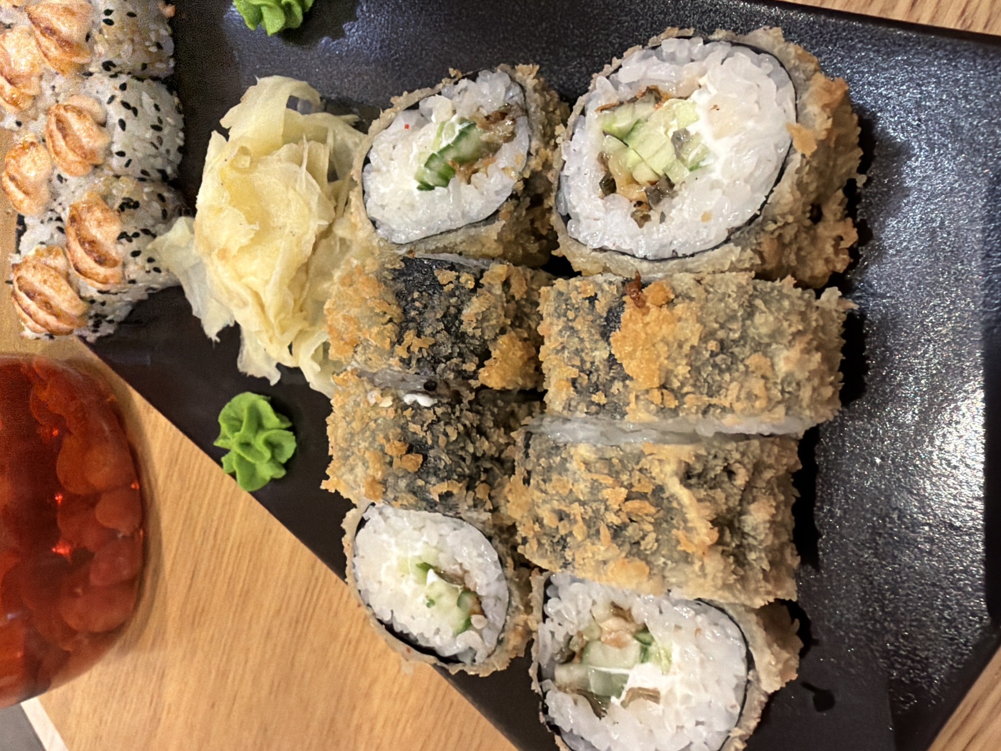 A black plate of sushi is set on a wooden surface. The sushi includes several pieces of deep-fried rolls with visible rice, seaweed, and creamy white filling with cucumber. There are also some slices of what appears to be pickled ginger and a dollop of green wasabi. In the upper left corner, a clear glass jar filled with red berries is visible. The lighting suggests an indoor setting, possibly a restaurant. The scene is captured from a slightly elevated, overhead perspective.