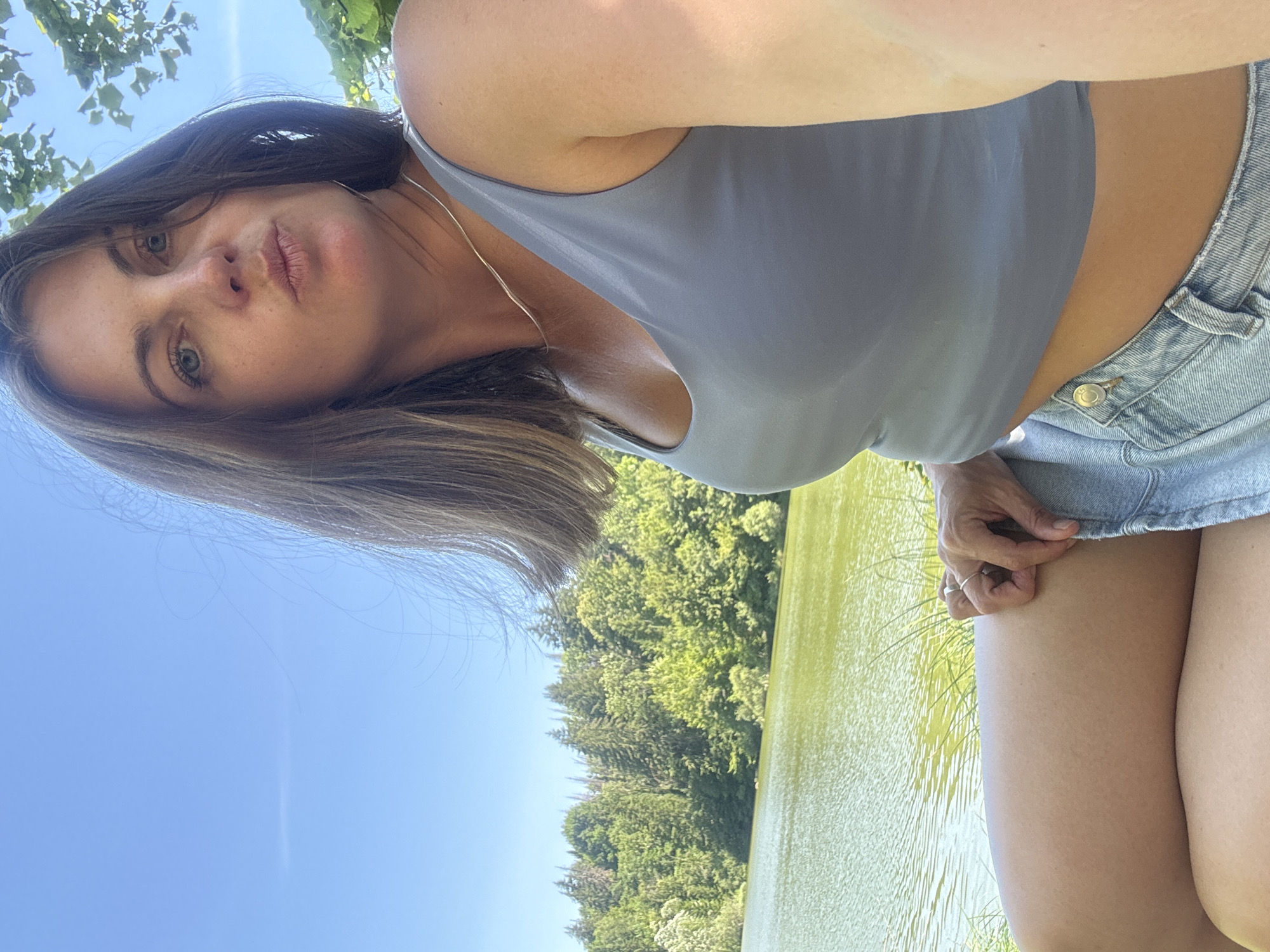 A woman with brown hair, wearing a grey tank top and denim shorts, is sitting outdoors by a lake. The lake water is green and reflects the clear blue sky. Behind the lake, a dense forest of green trees covers a hillside. The woman is looking at the camera with her lips pursed. A thin silver necklace is visible around her neck, and a ring is on her finger. The image appears to be taken on a sunny day.