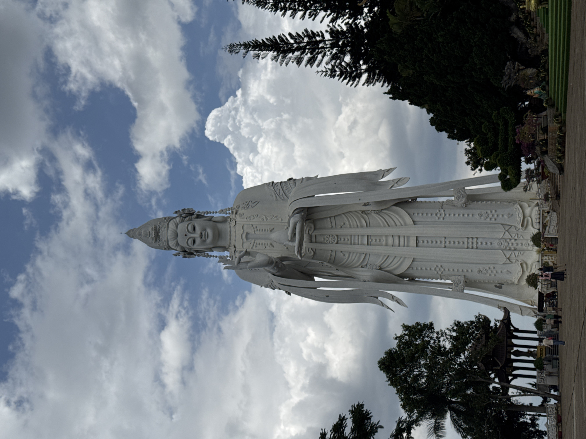 The image depicts a tall, white stone statue of a bodhisattva against a cloudy sky. The statue is situated outdoors, likely in a temple complex or park. The location is in the city of Nam Ban, Vietnam. Details of the statue's robes and headdress are visible, and it appears to be holding a vessel or object. A tree and a glimpse of a building appear in the bottom portion of the image, alongside a path and some people. The surrounding environment includes trees and vegetation.