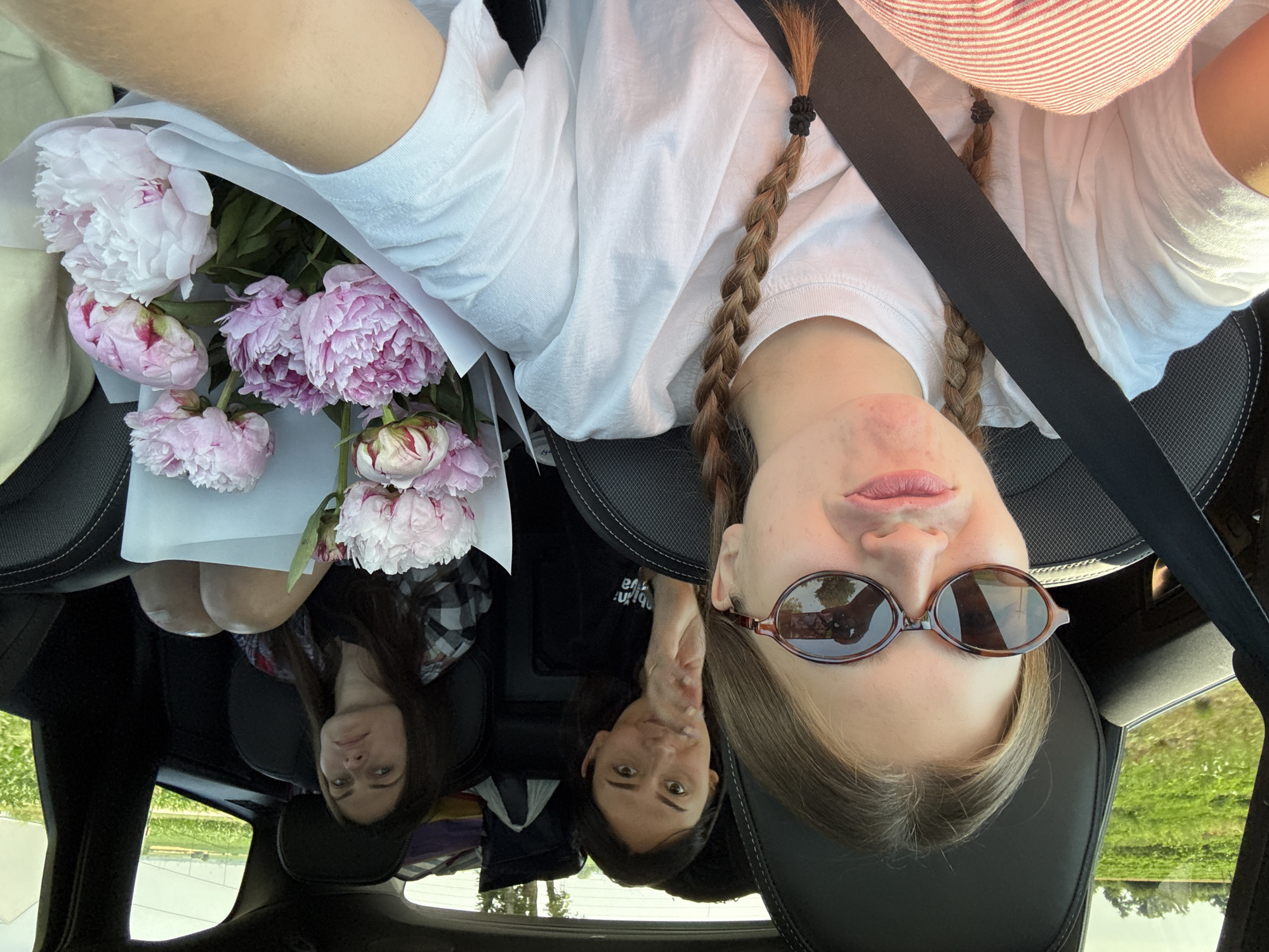 In this photograph, three young women are seated inside a car, possibly in Mandjelos, Serbia, given the rural landscape visible through the windows. The woman in the foreground, facing the camera, is wearing sunglasses and has her hair in braids. She is making a pouting gesture. To her right, a woman in the middle seat is looking forward and touching her chin. The third woman, in the back seat on the right, is looking away from the camera with a neutral expression. A bouquet of pink peonies is held between the two women in the front seats. The car's interior is dimly lit, with natural light filtering in from the outside, illuminating the green fields and trees. The women appear to be on a journey.