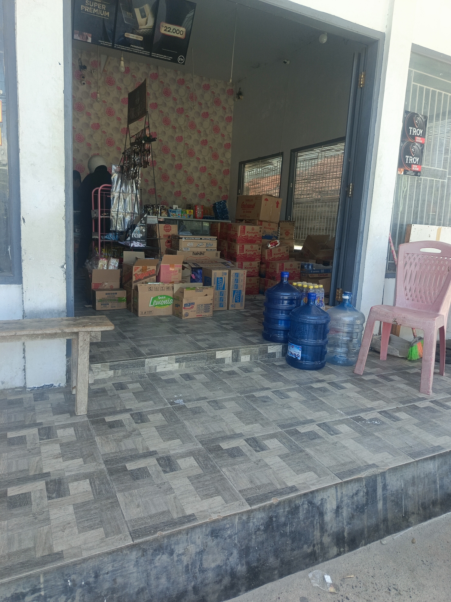 The image shows the exterior of a small convenience store in Tenggela, Indonesia. In the foreground, a grey tiled floor with a geometric pattern is visible, leading up to a raised concrete step. To the left, a simple wooden bench with two legs sits on the tiles.
The store entrance is open, revealing a cluttered interior. Boxes of various goods are stacked inside and in front of the entrance. Among the visible items are boxes labeled "Kompas," "Super," and "Lemonia." Several large blue water jugs are placed on the tiled floor just outside the entrance, with one clear jug also present.
To the right of the entrance, a pink plastic chair is positioned. Behind the chair, a broom with green and orange bristles leans against the wall. On the wall to the right of the entrance, two circular posters with the word "TROY" are displayed.
Inside the store, a person wearing a dark jacket and a white helmet is partially visible, appearing to be working or attending to something. A metal shelf unit holding more products is also seen within the store. The interior walls are decorated with a floral wallpaper featuring pink roses.
The overall scene suggests a typical small, local business in Indonesia, with a focus on selling various household goods and beverages. The presence of water jugs indicates they are likely a distributor or seller of bottled water.
