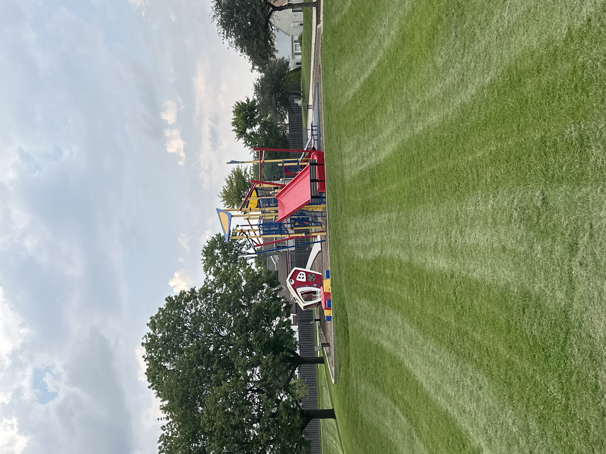 This is a full-shot photograph of a playground on a cloudy day. The playground equipment is centrally located and includes a red slide, a climbing structure with blue and yellow accents, and a small red playhouse with a white window. The playground is surrounded by a dark fence. To the left of the playground is a large tree with green leaves. In the foreground, a neatly mowed green lawn with visible mowing stripes stretches towards the viewer. In the background, residential homes with light-colored siding are visible. The sky is overcast with gray clouds and some patches of lighter sky. The scene appears to be set in a suburban neighborhood in Schaumburg, Illinois, USA. There are no people visible in the image.