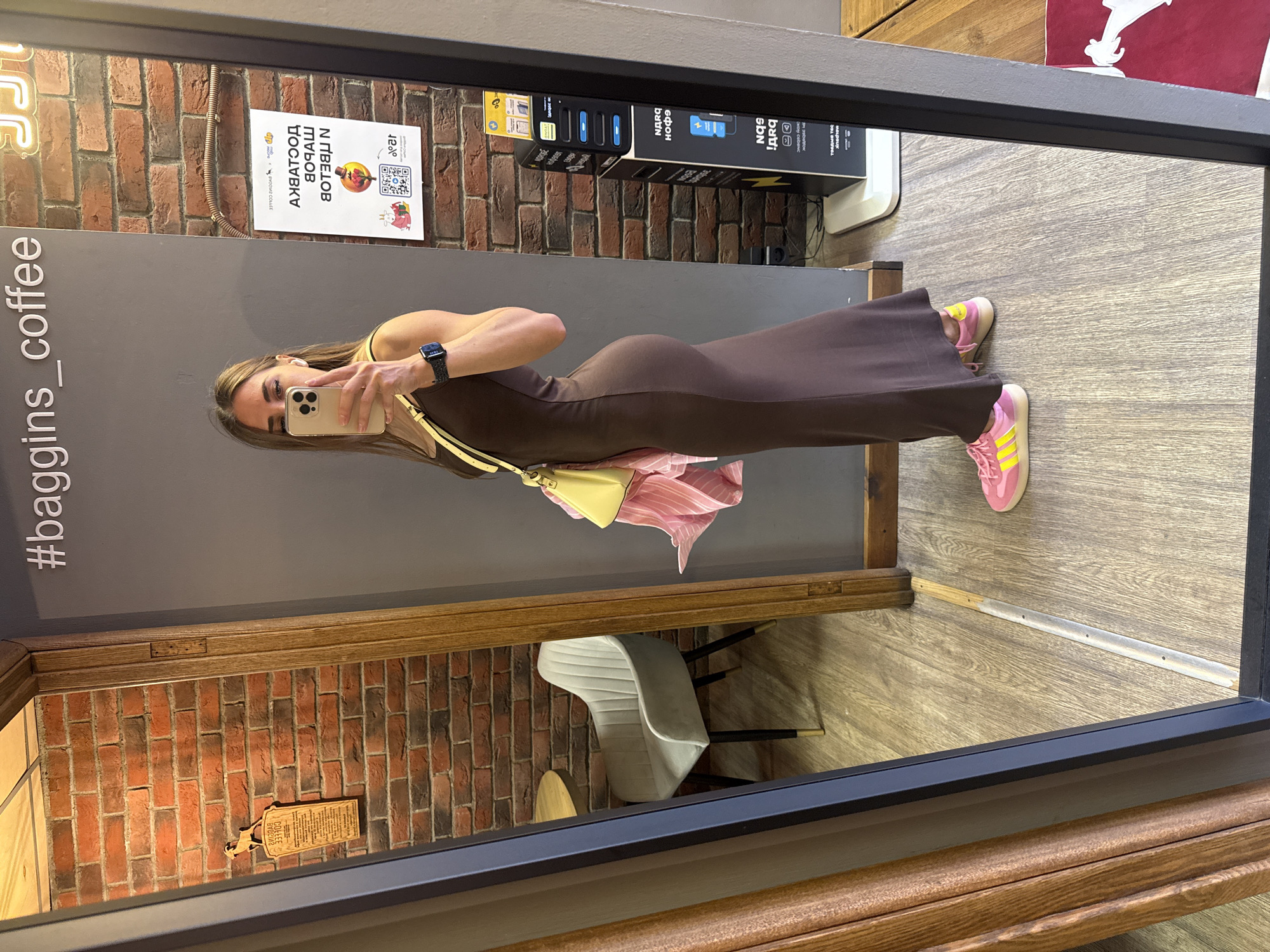 A woman is taking a mirror selfie in what appears to be a cafe, likely in Saint Petersburg, Russia. She is wearing a brown, form-fitting, floor-length dress and bright pink and yellow sneakers. She has a yellow purse slung over her shoulder and is wearing a smartwatch on her left wrist. The background of the mirror shows a brick wall, a grey wall with text that reads "#baggins_coffee", a light-colored chair, and a wooden table. A white sign with Cyrillic text and a QR code is also visible on the brick wall behind her. The floor is made of light-colored wood planks. The overall lighting suggests an indoor environment.