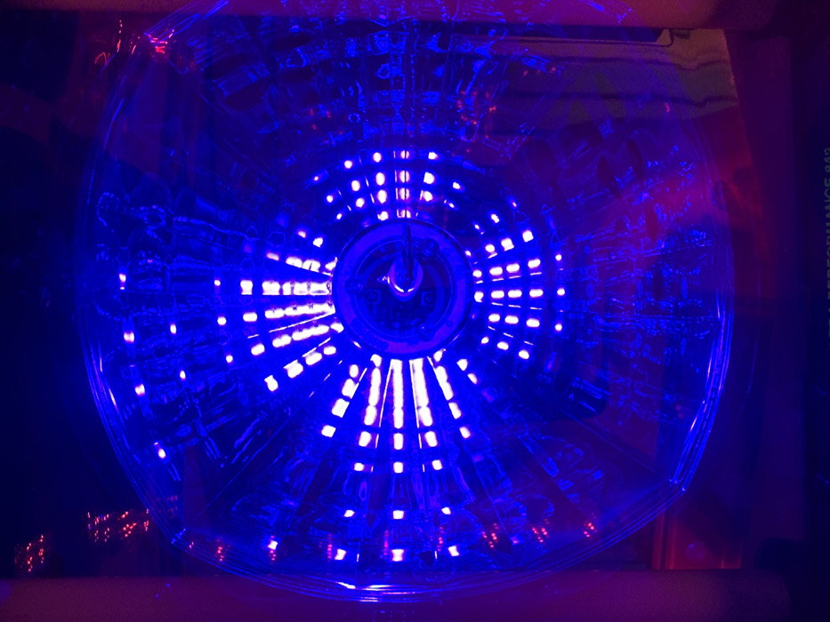 A circular, transparent object with embedded blue lights is captured in a dark environment. The lights are arranged in concentric rings, radiating outward from the center. Reflections and distortions create a blurry, dynamic effect. Hints of red light are noticeable in the lower left and right corners. The words "ULTRA PERFORMANCE 942" are partially visible on the right side.