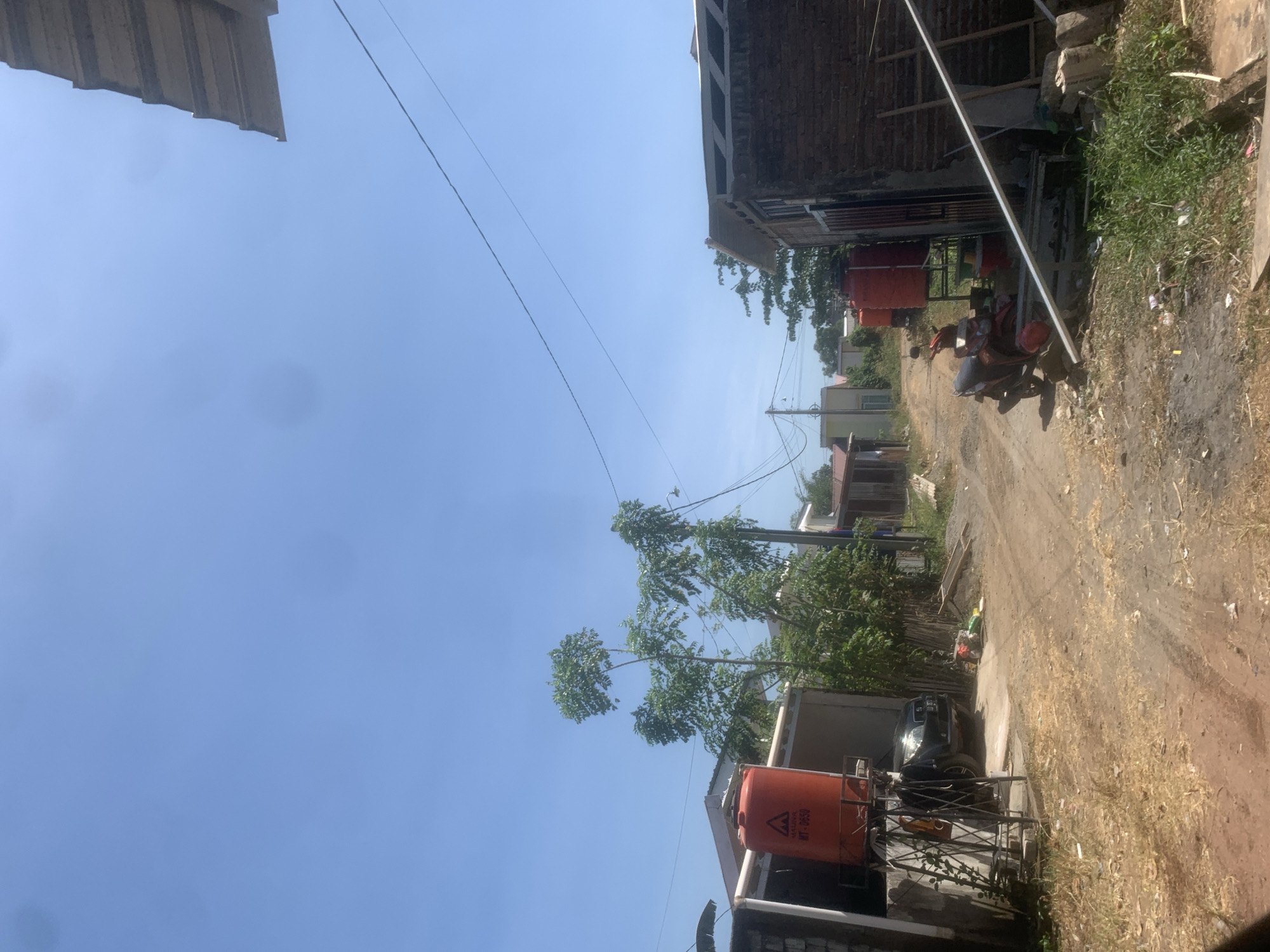The image depicts a street scene in Bonto Bunga, Indonesia. The foreground is a dirt road with patches of dry grass and scattered debris. To the left, a black SUV is parked partially under a structure with a large orange water tank on a metal stand. Further to the left, a low concrete wall and a building are visible.

In the middle ground, the dirt road continues, flanked by vegetation and several buildings. A red scooter is parked on the right side of the road, near another orange water tank. A tall, thin tree with lush green foliage stands prominently in the center.

In the background, more buildings and trees can be seen under a clear blue sky with some power lines crisscrossing the frame. The overall impression is of a somewhat rural or developing urban area. There are no people visible in the image.