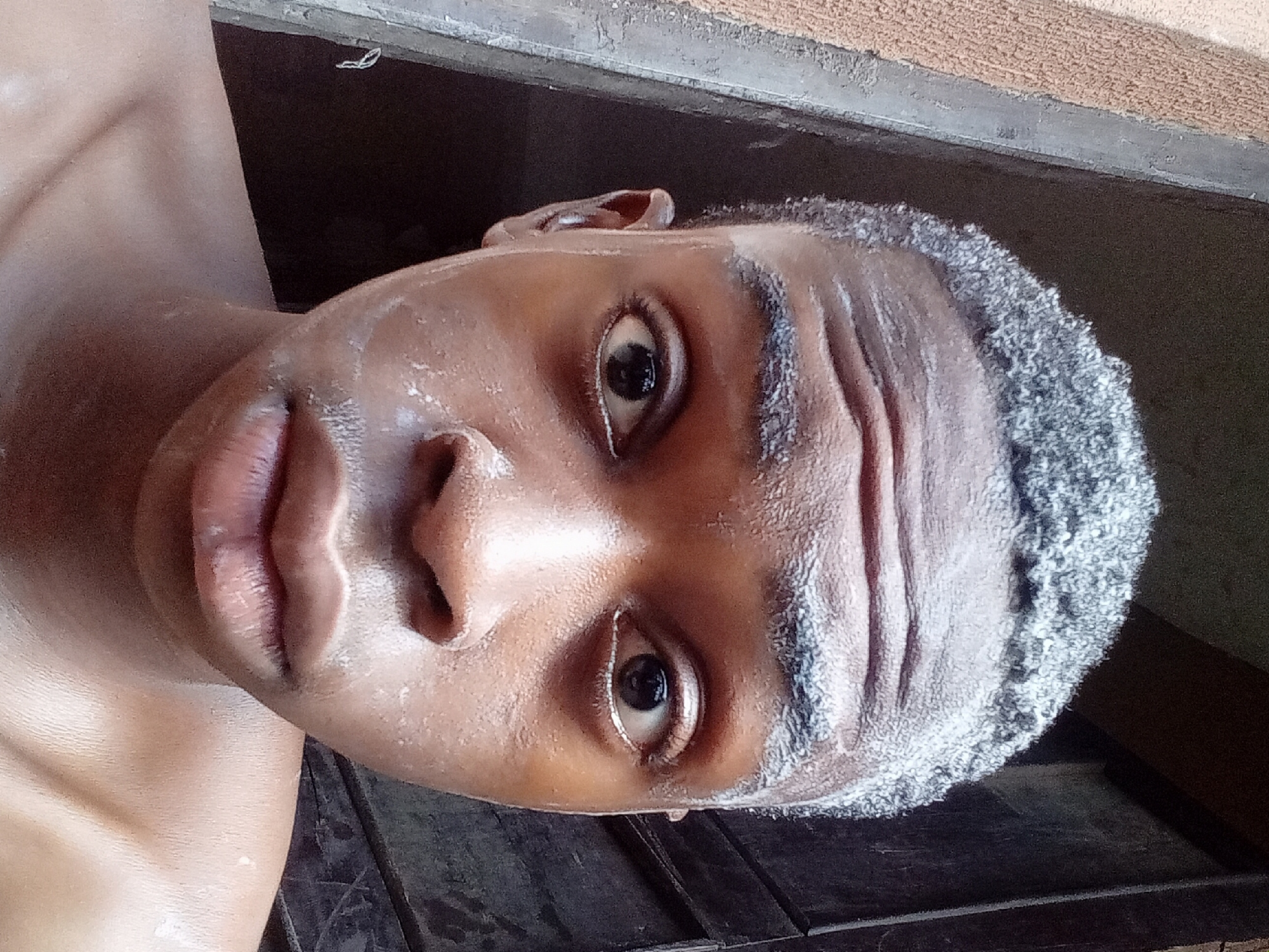 A close-up, high-angle, rotated view shows the face and upper torso of a Black male. His skin is shiny, and his hair is short and curly with graying at the hairline. He has dark eyes and full lips. The background includes a dark, wooden surface and a tan wall with a dark, possibly shadowed, area above. It is not possible to identify the location as Onitsha, Nigeria, or provide further details without additional context.