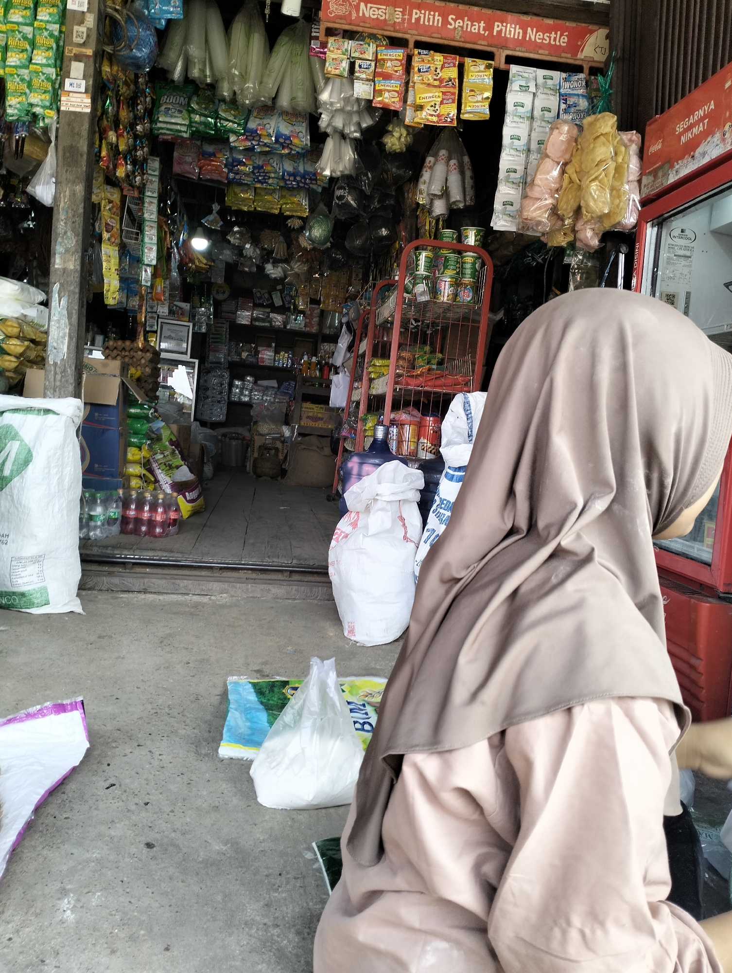 The image shows a street-level view of a small convenience store, possibly located in the city of Serdang Jaya, Indonesia. The store appears to be stocked with a variety of goods, including packaged food items, drinks, and household supplies, displayed on shelves and hanging from the ceiling. A person wearing a hijab is partially visible in the foreground, facing away from the camera and towards the store entrance. The ground in front of the store is made of concrete and shows some debris. The overall scene suggests a typical local market or shop in Indonesia.