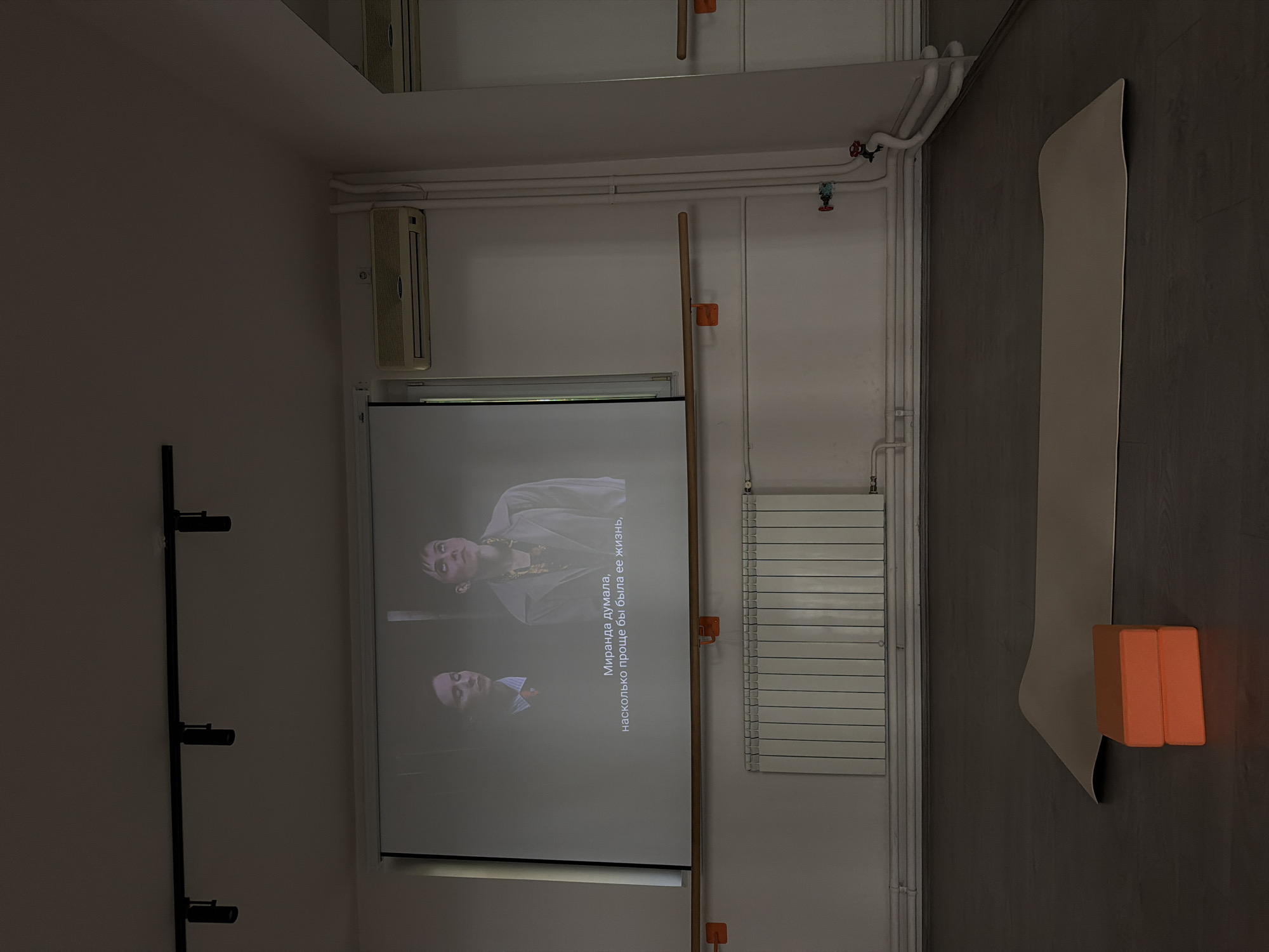 The image depicts an indoor space, likely a dance studio or fitness room, with a projector screen displaying two women. The screen has Cyrillic subtitles that read "Миранда думала, насколько проще бы была ее жизнь," which translates to "Miranda thought, how much simpler her life would be." In the foreground, on the dark wooden floor, lies a light-colored yoga mat and two stacked orange yoga blocks. The room features white walls, a white radiator underneath the projector screen, and overhead track lighting. An air conditioning unit is visible on the wall. The overall lighting is dim, with the projector screen being the primary light source. There are no discernable geographic markers to place this scene in Vracar Urban Municipality, Serbia.