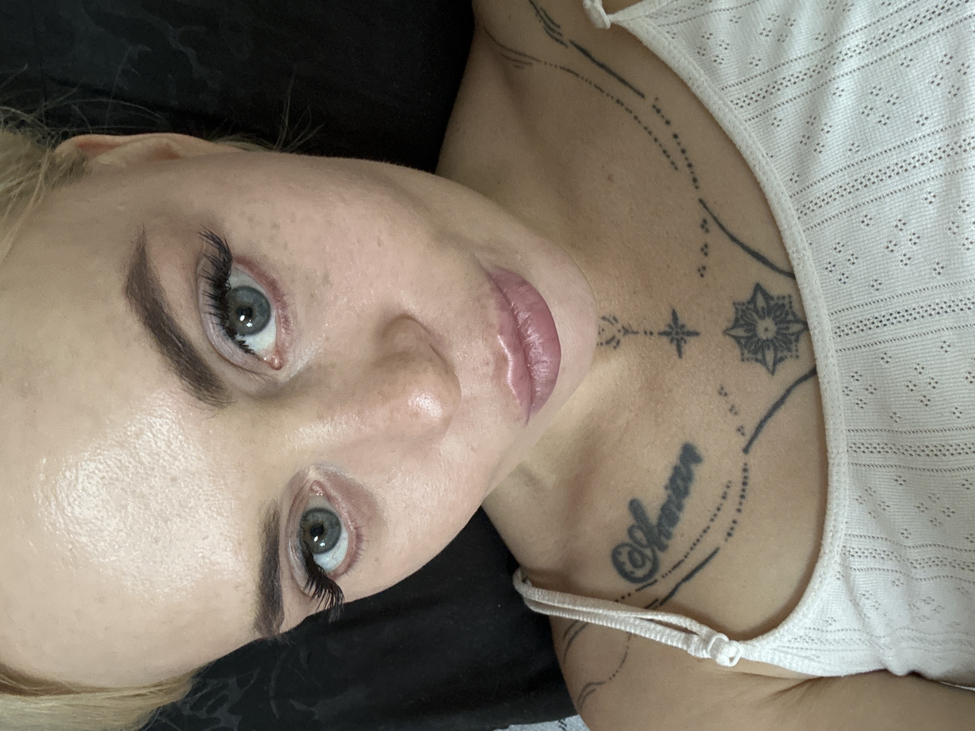 A close-up, high-angle shot shows a woman with blonde hair, light skin, and blue eyes. She is wearing a white, ribbed tank top with thin straps. The woman has a tattoo on her chest that appears to read "Aman" in a cursive font, along with other decorative tattoo elements. Her makeup includes eyeliner and mascara, accentuating her eyes. Her lips are slightly parted and appear to have a light pink lip gloss. The background is dark and out of focus.