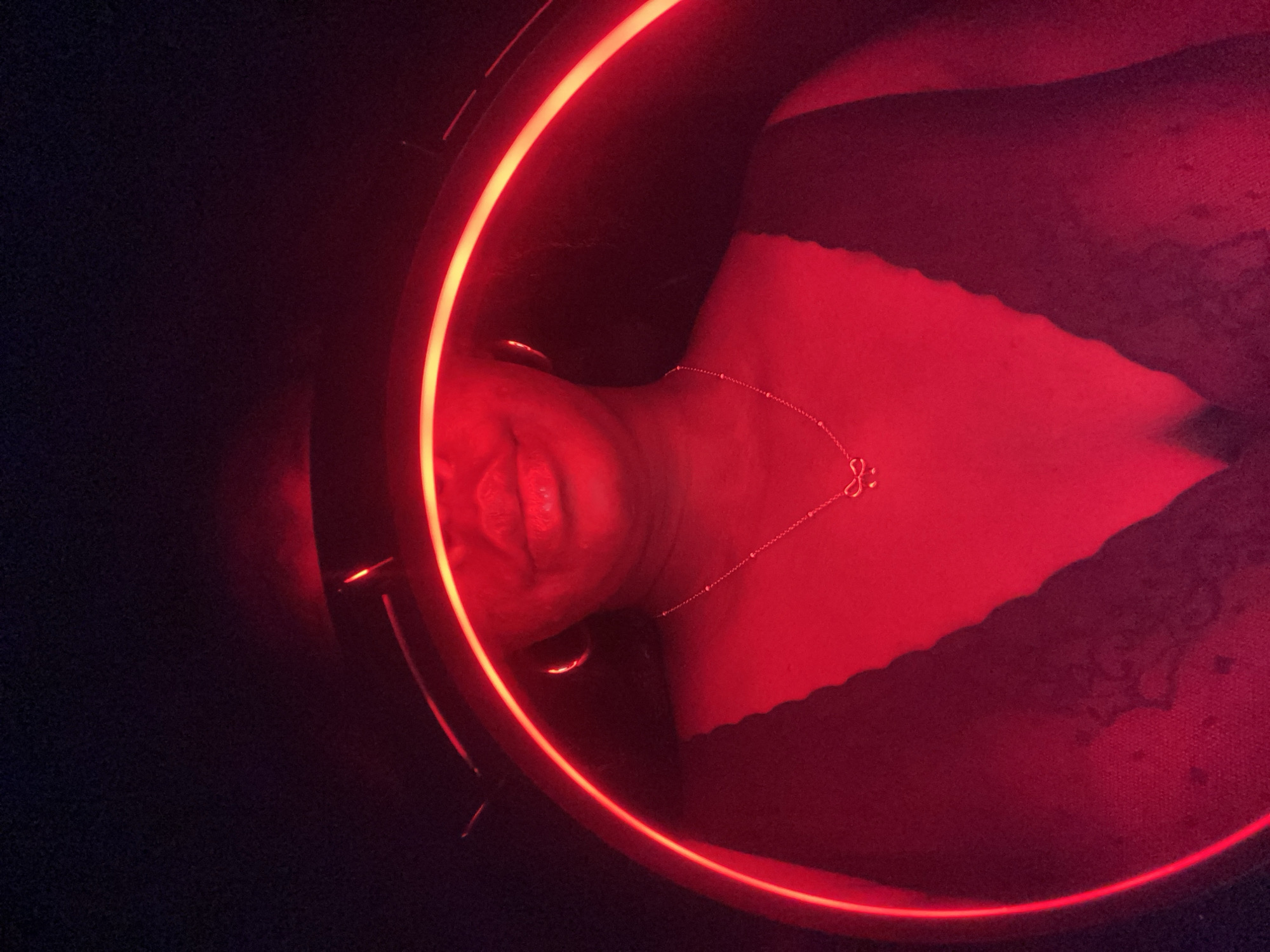 A close-up image shows a person's face and upper chest illuminated by a red neon ring. The person is wearing a necklace. A dark fabric is visible at the top of the frame. The location is not discernible.