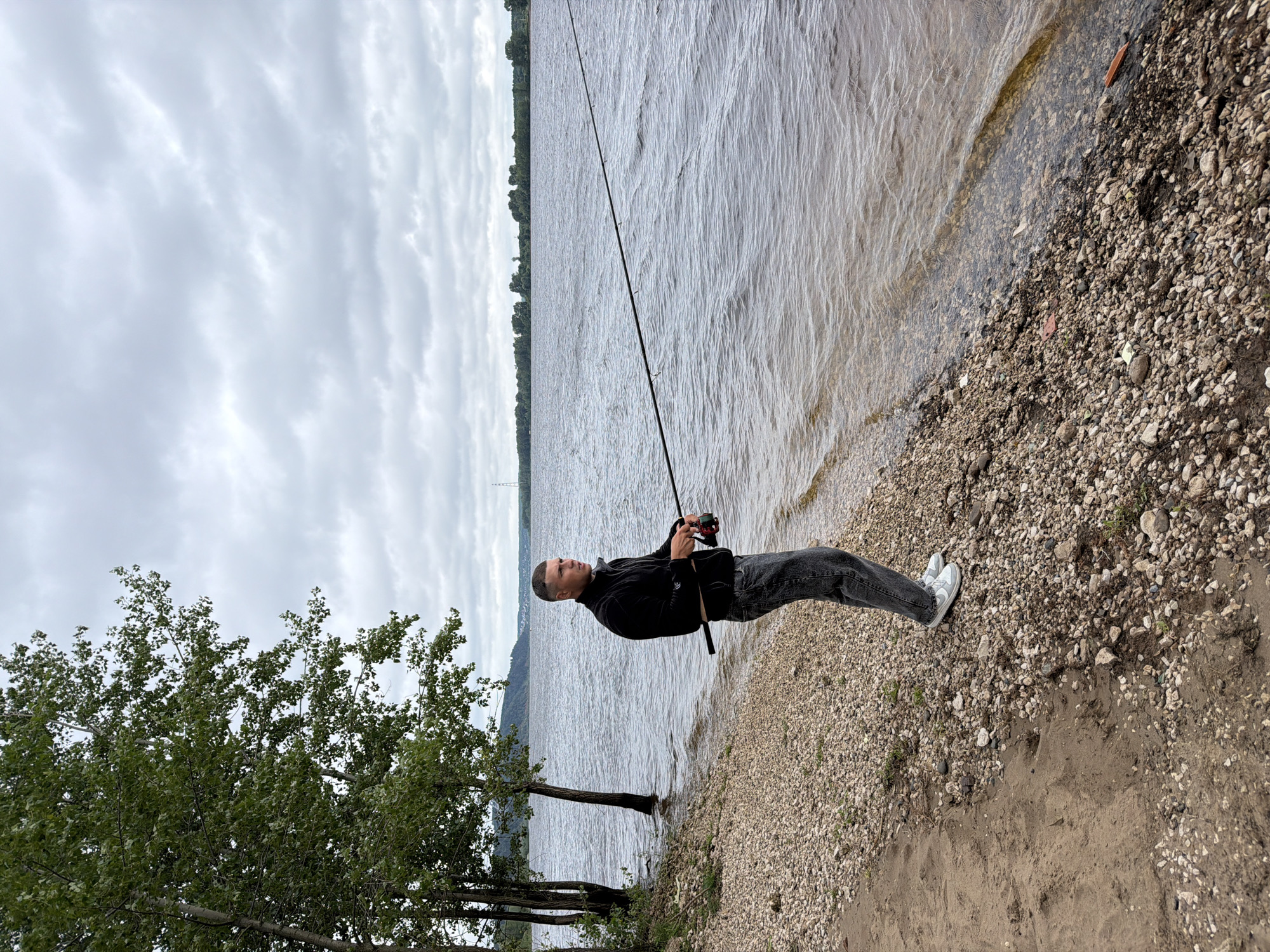 A man stands on the pebbly shore of a wide river, fishing. He is wearing a black jacket, grey jeans, and white sneakers. The man is holding a fishing rod, with the line cast into the water. The river is calm, with a slight ripple on the surface. In the background, a treeline stretches across the horizon, with a few distant structures visible. The sky is overcast and cloudy, suggesting a cool, possibly humid day. The specific location is along the Kama River in the city of Chaikovsky, Russia, near the embankment.