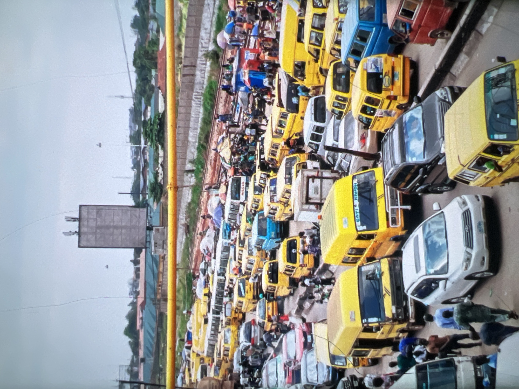 This is a high-angle, long shot of a congested street in Lagos, Nigeria. The scene is dominated by a dense traffic jam of various vehicles, predominantly yellow minibuses, commonly known as danfos, a staple of Lagos public transportation. Several other vehicles, including cars, SUVs, and a few trucks, are also visible, interspersed amongst the danfos. The road appears to be a multi-lane thoroughfare, heavily packed with vehicles, indicating a significant traffic standstill.
Pedestrians are scattered throughout the scene, navigating between the stationary vehicles. Some are standing in groups, others appear to be walking, and a few are in proximity to what seem to be market stalls or informal vendor setups, indicated by colorful umbrellas and awnings. The presence of these stalls suggests a busy commercial area or a transport hub.
In the background, a large, rectangular building with a distinctive lattice-like facade stands tall, possibly a commercial or administrative building. Beyond this structure, there are more buildings and greenery, suggesting an urban environment with some developed infrastructure. The sky is overcast, contributing to a muted lighting of the scene. The overall impression is one of a bustling, chaotic, and densely populated urban environment in Lagos, characterized by heavy traffic and a vibrant street life.