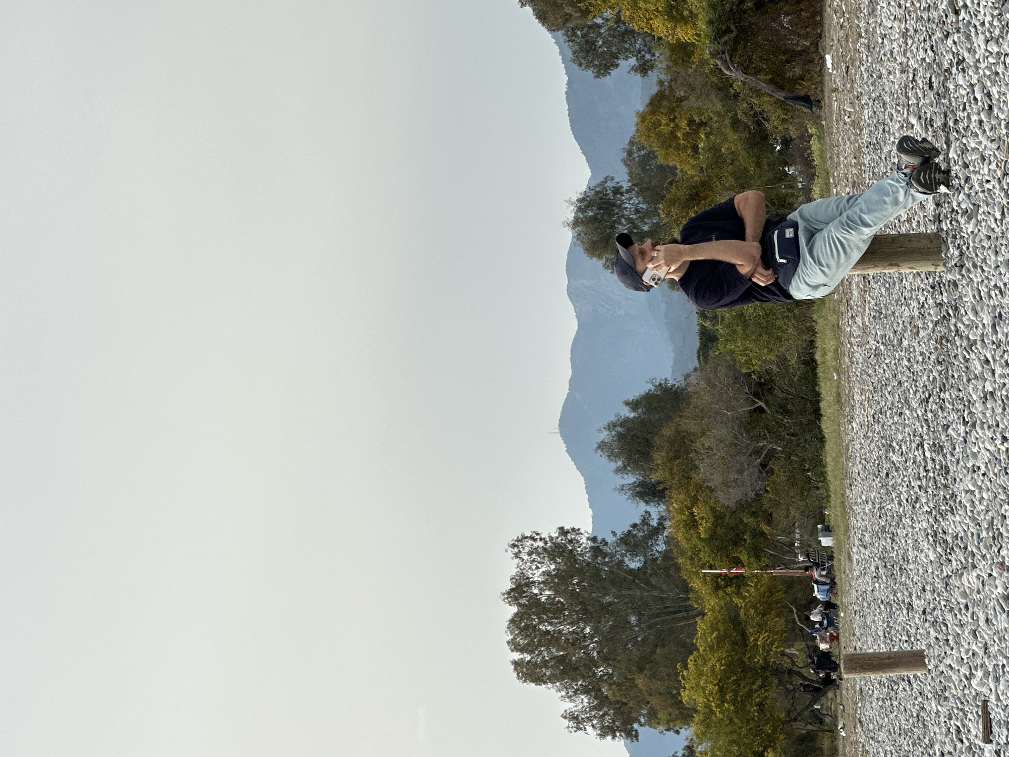 A man is sitting on a wooden post on a ground covered with small rocks. He is wearing a dark blue shirt, light blue pants, a dark cap, and black-and-white sneakers. He's holding a white phone to his ear, seemingly engaged in a conversation. A black fanny pack is strapped around his waist.
In the background, there are trees with green and yellow foliage, partially obscuring a mountain range. Further back, a gathering of people is noticeable. The sky is overcast.
The location is likely somewhere near the coast in Fethiye, Turkey, given the combination of rocky ground, trees, mountains, and the presence of tourists.