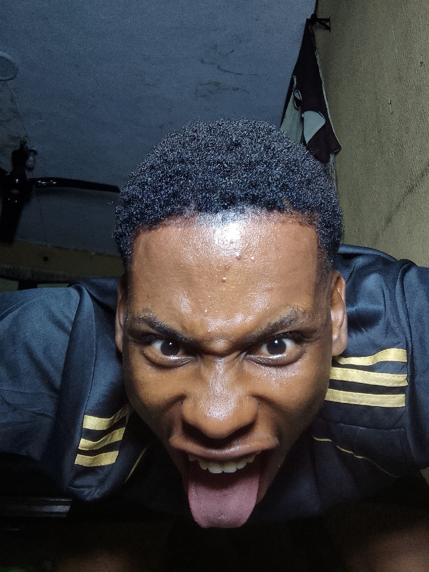 A close-up, low-angle shot shows a man's face with an intense expression, sticking his tongue out. His skin is dark and appears sweaty, and he has short, dark curly hair. He is wearing a black shirt with gold stripes on the sleeves. Behind him, the wall is yellowish and there is a white and black object hanging on it. A ceiling fan is partially visible at the upper left. The location and city are Unknown.