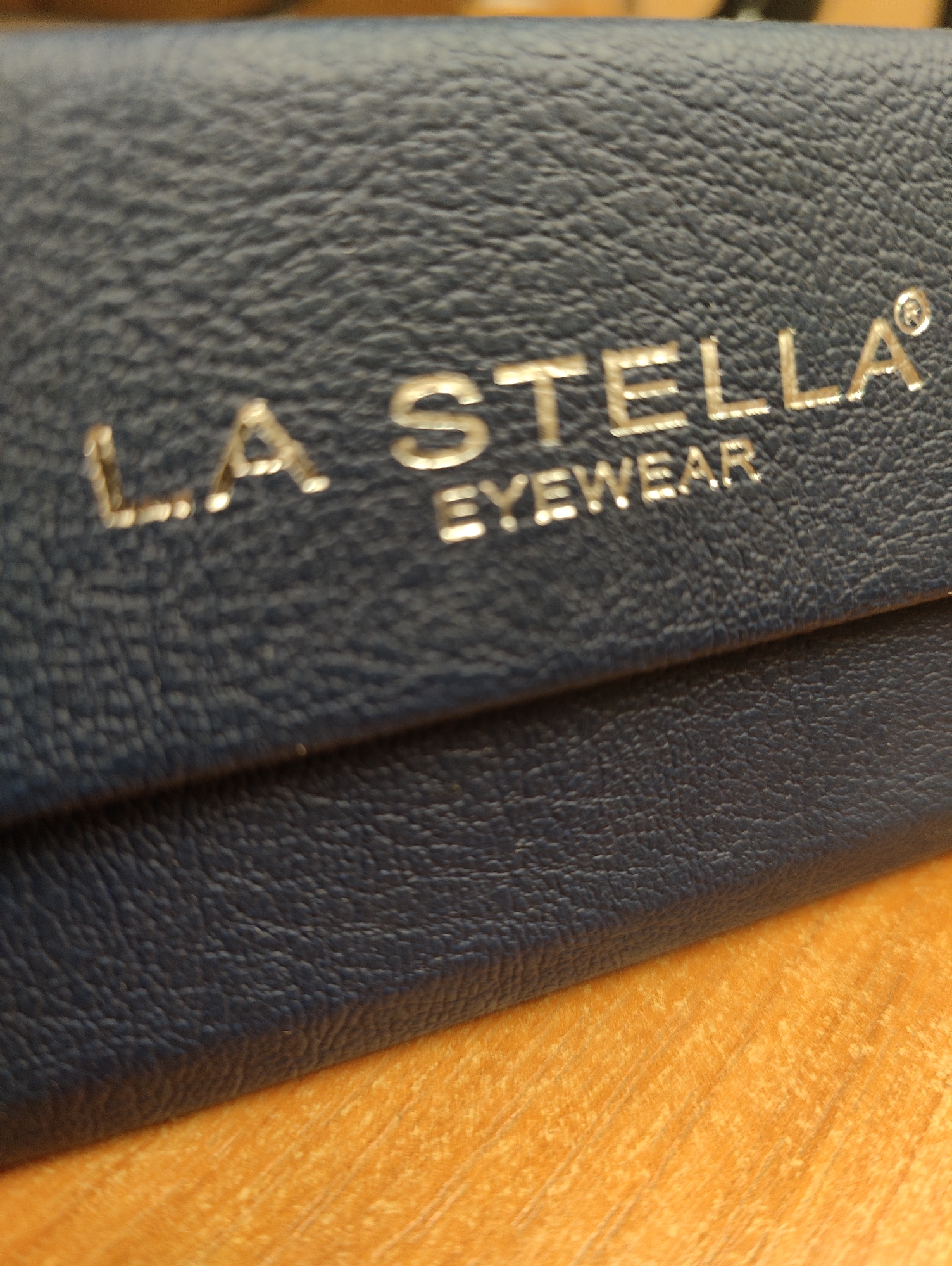 The image shows a close-up, slightly blurred view of a dark blue, textured material, likely leather, with gold lettering embossed on it. The lettering reads "LA STELLA" in larger font on the top line and "EYEWEAR" in smaller font directly below. The material appears to be a case or cover for eyewear. The bottom portion of the image reveals a bright orange, textured surface, possibly a wooden table or countertop, with visible grain patterns. The lighting is direct, creating some glare on the gold lettering. The overall scene is an indoor still life with a focus on product detail. The location is unknown.