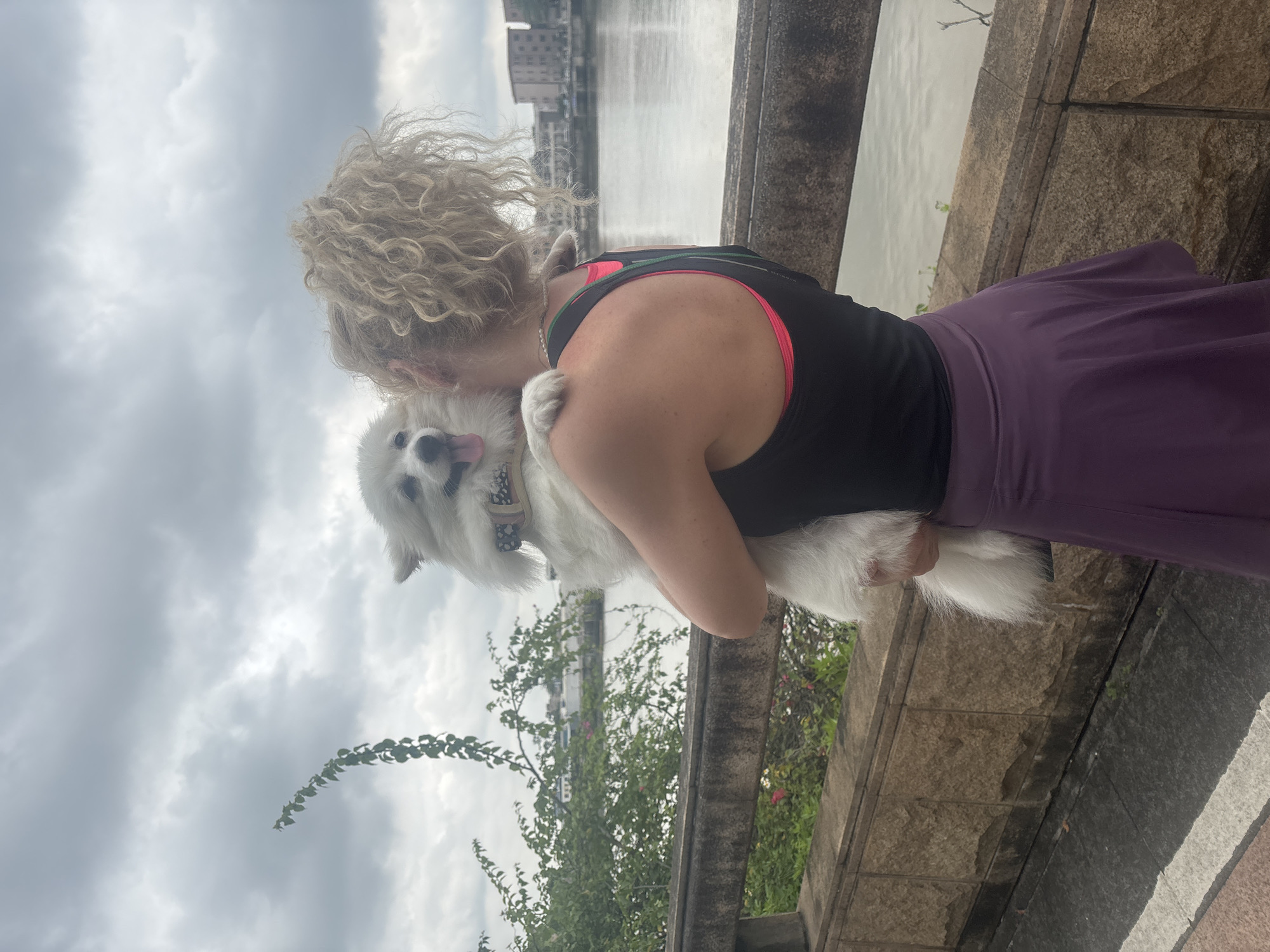 A woman with curly blonde hair holds a white fluffy dog in her arms, facing away from the camera. The dog has its tongue out and appears happy. The woman is wearing a black tank top with a pink sports bra underneath and a purple skirt. They are standing on a pathway next to a stone wall. In the background, there is a body of water, possibly a river, with buildings visible on the opposite bank, consistent with the Nanhai District in China. The sky is overcast with gray clouds. Green foliage is visible on the left side of the frame.