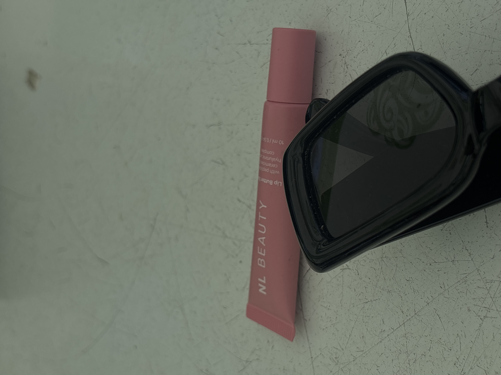 A pink tube of NL Beauty Lip Butter lies on a light-colored, textured surface, possibly a desk or countertop. The tube is angled, with the brand name "NL BEAUTY" clearly visible in white lettering. Text on the tube also indicates "Lip Butter," "with peptides," "ceramides," "hyaluronic complex," and "10 ml / 0.34 fl. oz." Partially obscuring the lower portion of the tube is a black object, appearing to be a pair of sunglasses or a compact mirror with a black frame. The surface has numerous fine scratches and some faint smudges. The background is a plain, light green hue with subtle variations in shading. There is no indication of the location within the city of Varna, Bulgaria.