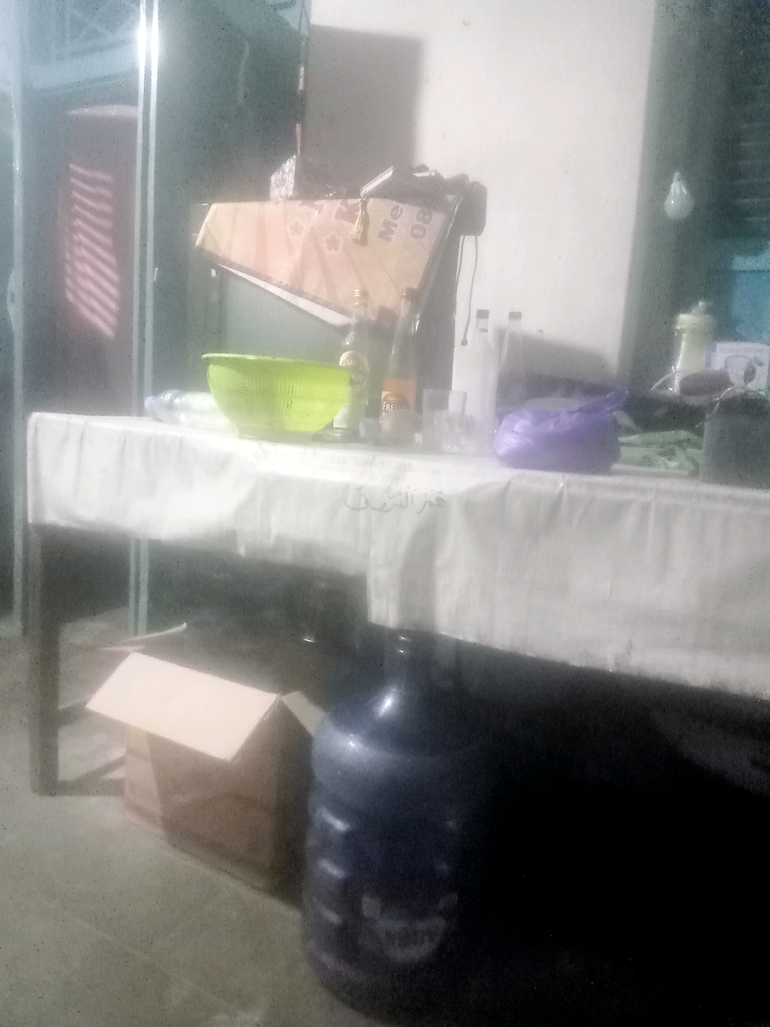 The image shows a cluttered indoor scene, possibly a storage or preparation area, located in Dukuhlo, Indonesia. In the foreground, a large, dark blue water jug is partially visible on the right. To the left of the jug, an open cardboard box is situated on the floor. The middle ground features a table covered with a white tablecloth, on which several items are placed. These include a bright green colander, several bottles, and a purple plastic bag. A patterned box with tassels is propped up behind the bottles. The background is out of focus, but appears to show shelves and more storage units. The overall lighting is dim, with some light sources creating reflections and glare on the surfaces.