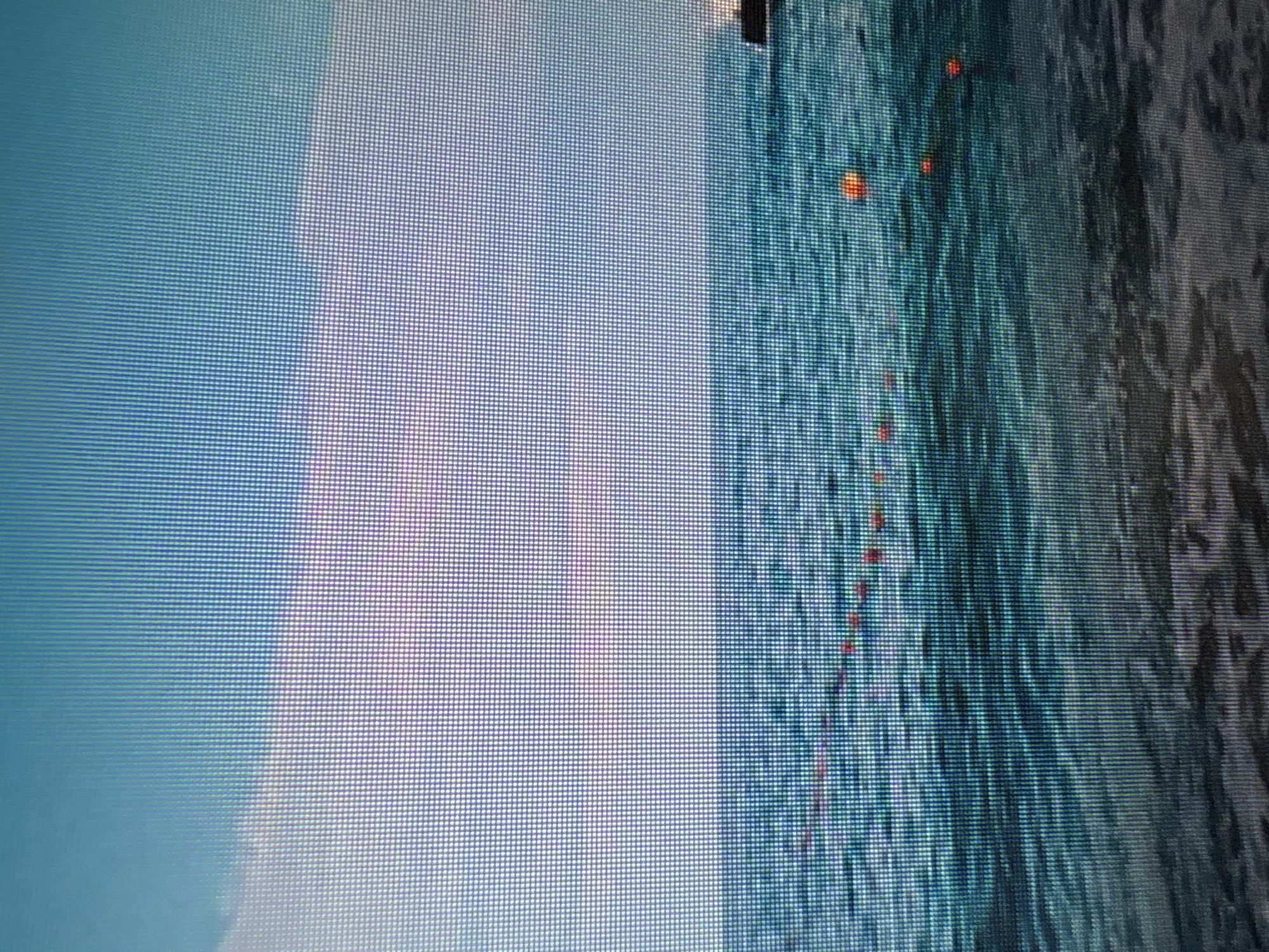 The image is a photograph of a body of water, likely the sea or a large lake, under a bright sky with some clouds. The water is a deep blue-green, with gentle waves visible. A line of orange buoys is floating on the surface, extending into the distance. In the far right of the image, a dark structure, possibly a pier or a building, is partially visible. The foreground shows a sandy or rocky shoreline. The image quality is low, with visible pixelation, suggesting it might be a digital screenshot or a photograph taken from a screen. There are no people visible in the image. The location is not Moscow, Russia.