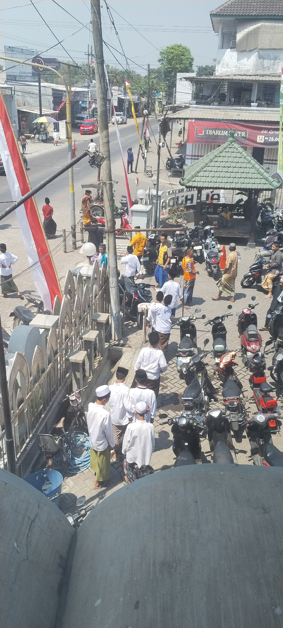 In the city of Jombang, Indonesia, on a sunny day, a street scene unfolds with numerous individuals, many dressed in traditional Islamic attire, congregating near a building with a green, pagoda-style roof. Several motorcycles are parked haphazardly on the paved ground. In the background, a red car is visible on the road, alongside other buildings and trees. The image appears to have been taken from an elevated position, looking down upon the bustling street. A flag, predominantly red and white, is draped over a railing in the foreground. The atmosphere suggests a community gathering or event, possibly related to religious activities.