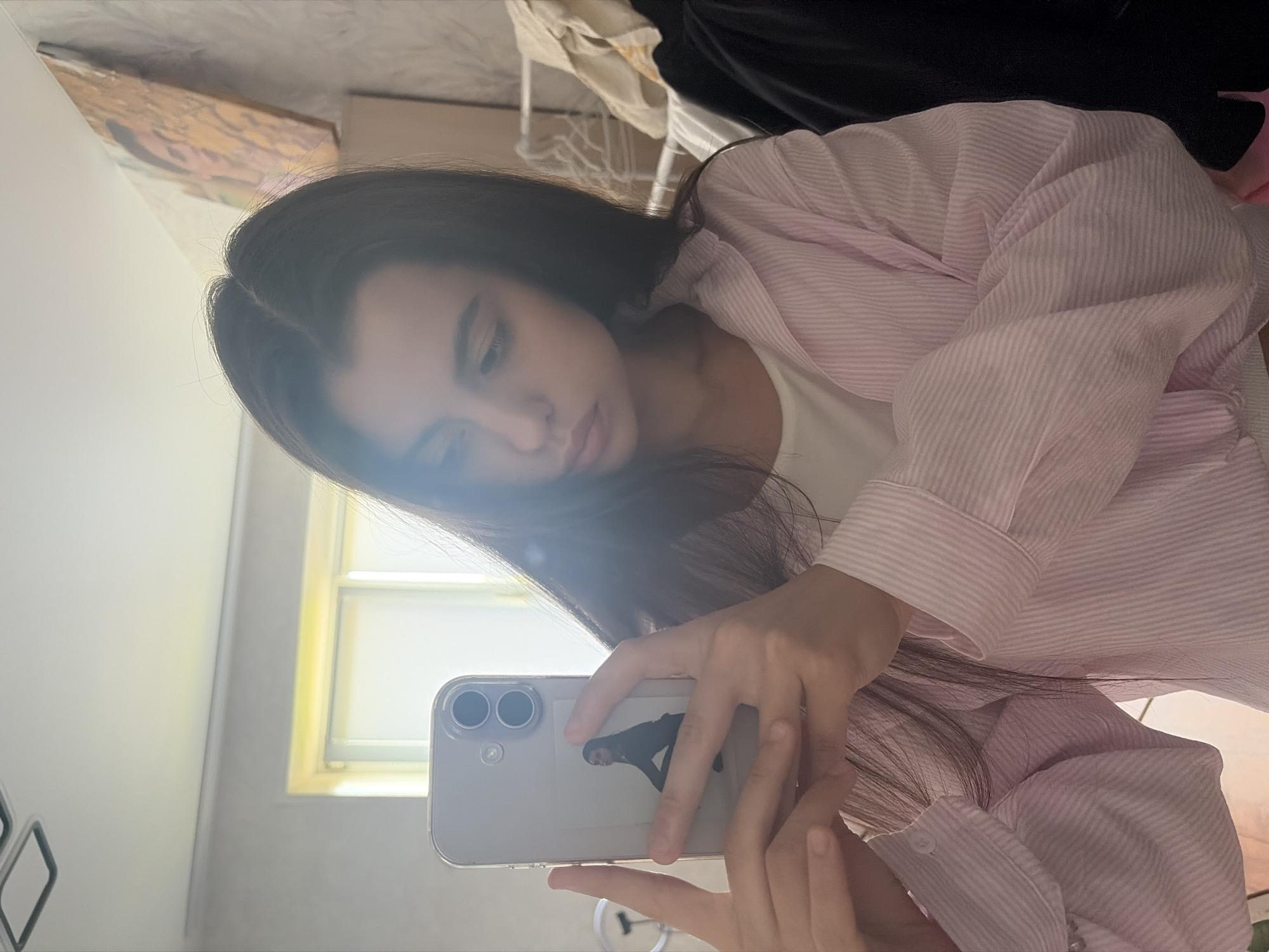 In an indoor setting, a young woman with long, dark hair takes a selfie in a mirror. She is wearing a light pink and white pinstripe button-down shirt over a white top. She holds a silver smartphone in her left hand, which displays a small sticker of a person in a dark outfit. Her right hand is visible, supporting the phone. The background suggests a room with natural light filtering in from a window, illuminating the scene. A painting with warm tones is visible on the wall to the right. The image does not contain any specific elements that indicate it was taken in Chaikovsky, Russia.