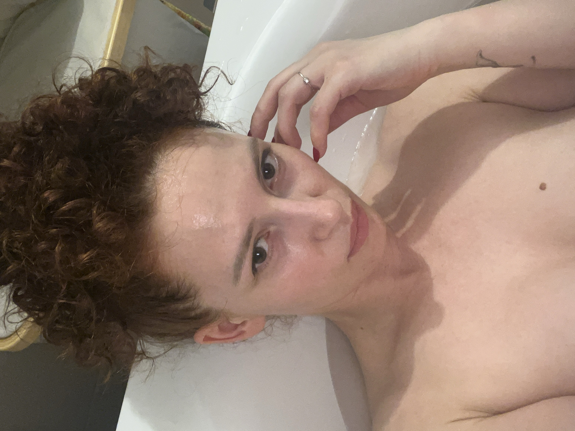 A woman is lying in a white bathtub. She has curly red hair and fair skin. She is wearing a ring on her left hand. There is a tattoo on her left arm. Her nails are painted red. She is looking at the camera. The location is not discernible.