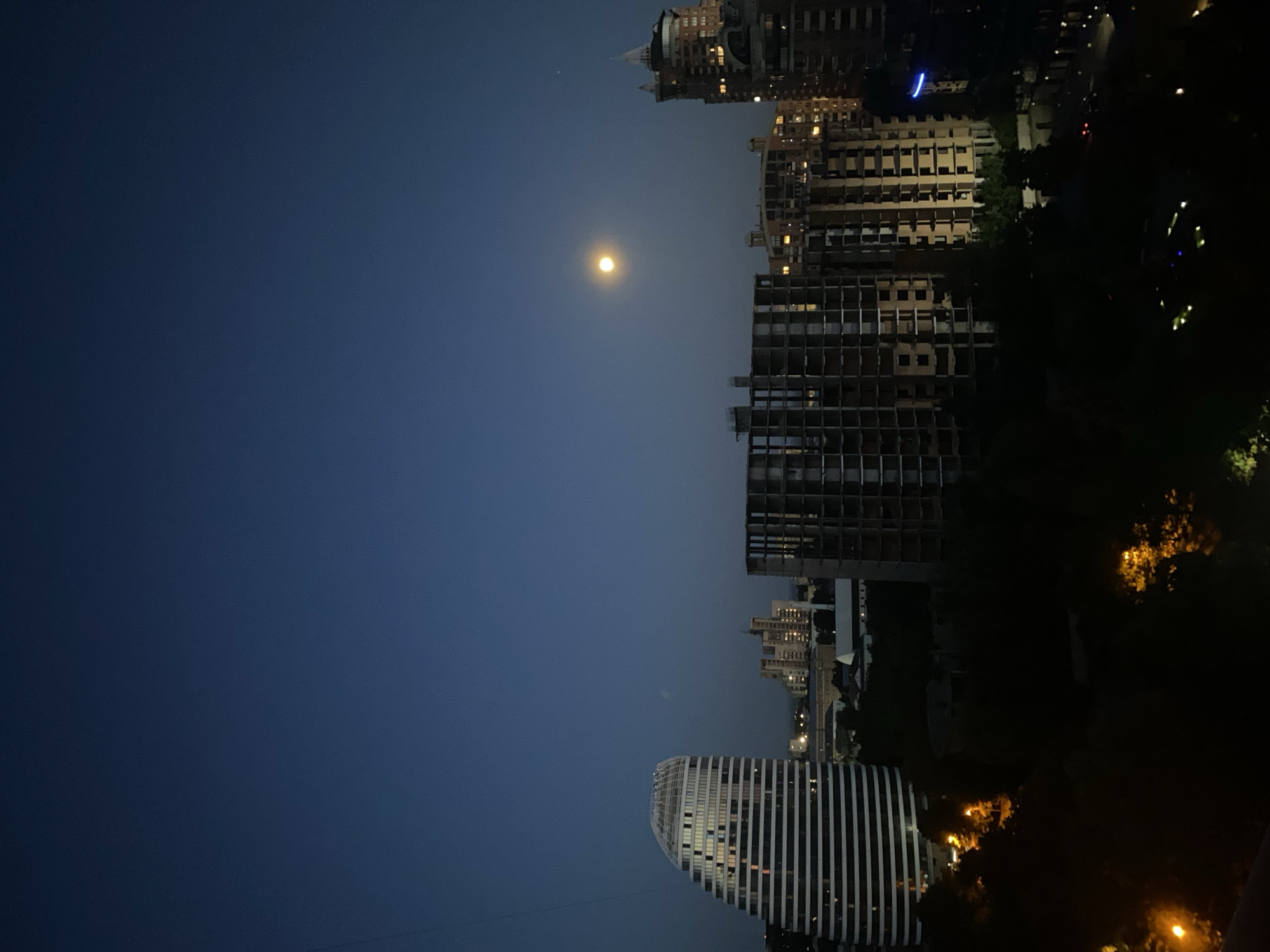 The image depicts a nighttime cityscape in Kyiv, Ukraine. In the foreground, dark foliage obscures the lower portion of the scene. Silhouetted trees with some illuminated branches are visible, suggesting the presence of streetlights or building lights filtering through.
Dominating the middle ground are several buildings. On the left, a distinctive, curved, white building with horizontal black stripes and illuminated windows stands prominently. To its right, a large, unfinished concrete building with visible floor plans and structural elements is under construction. Further to the right, another tall, multi-story building with illuminated windows and a conical roof section is visible. In the background, more urban structures and lights of the city are faintly discernible.
The sky is a deep, dark blue, characteristic of twilight or nighttime. A bright, full moon is positioned in the upper right quadrant of the sky, casting a soft glow. The overall impression is of an urban environment at dusk or night, with a mix of completed and under-construction buildings. The image appears to be taken from an elevated vantage point, possibly a rooftop or balcony.