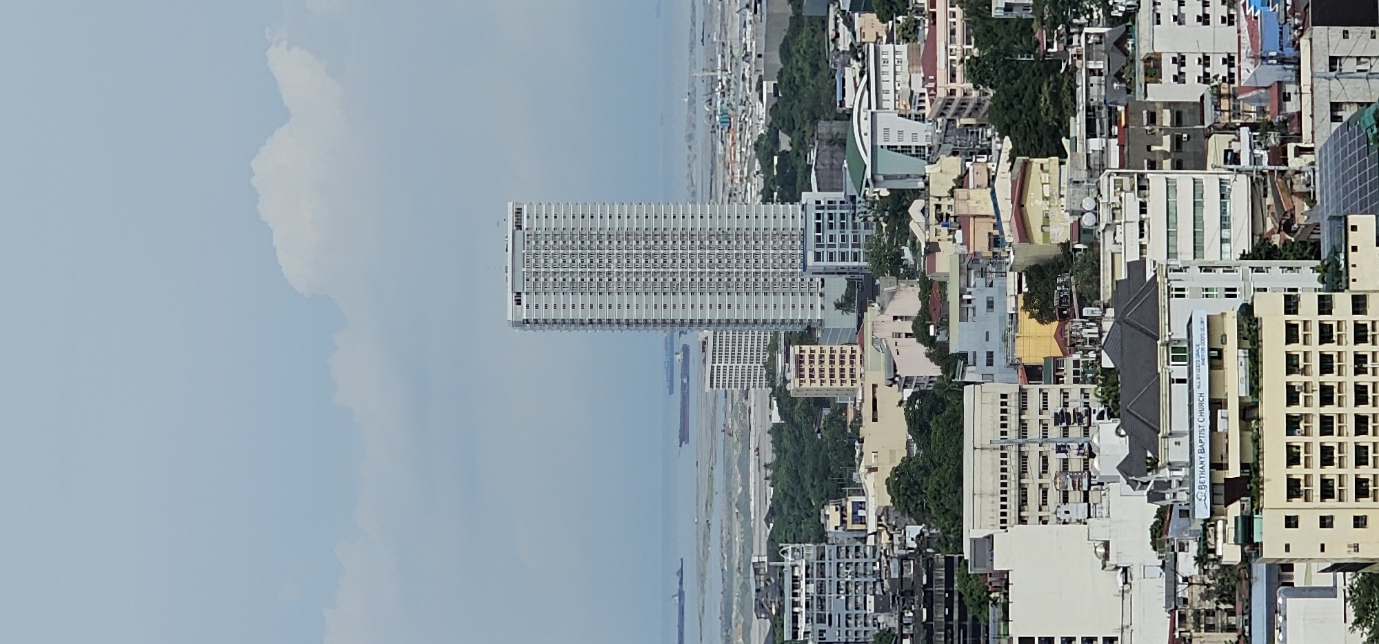 This is a medium-shot, eye-level, vertical photograph of a dense urban cityscape in Makati, Philippines, under a hazy, overcast sky with a few wispy clouds. The primary subject is a very tall, slender, light-colored apartment building dominating the center-right of the frame, its facade covered in rows of windows. In the distance, a body of water, likely Manila Bay, is visible with several cargo ships on the horizon.
The foreground and midground are filled with a mix of buildings of varying heights and architectural styles, indicative of a bustling urban environment. Many of these structures are residential or commercial buildings, featuring numerous windows, balconies, and different colored facades, ranging from white and beige to muted blues and yellows. Some buildings appear older with more traditional designs, while others are more modern. Lush green trees are interspersed among the buildings, suggesting parks or tree-lined streets within the city.
A prominent structure in the lower center of the image is a building with a sign that reads "BETHANY BAPTIST CHURCH" with the inscription "ALL BY GOD'S GRACE AND FOR GOD'S GLORY." This building has a distinct dark-colored roof with multiple peaks and appears to be a place of worship or community center. Solar panels are visible on the rooftop of a building in the bottom right corner. The overall impression is one of a densely populated city with a mix of residential, commercial, and religious structures, situated near a major body of water.