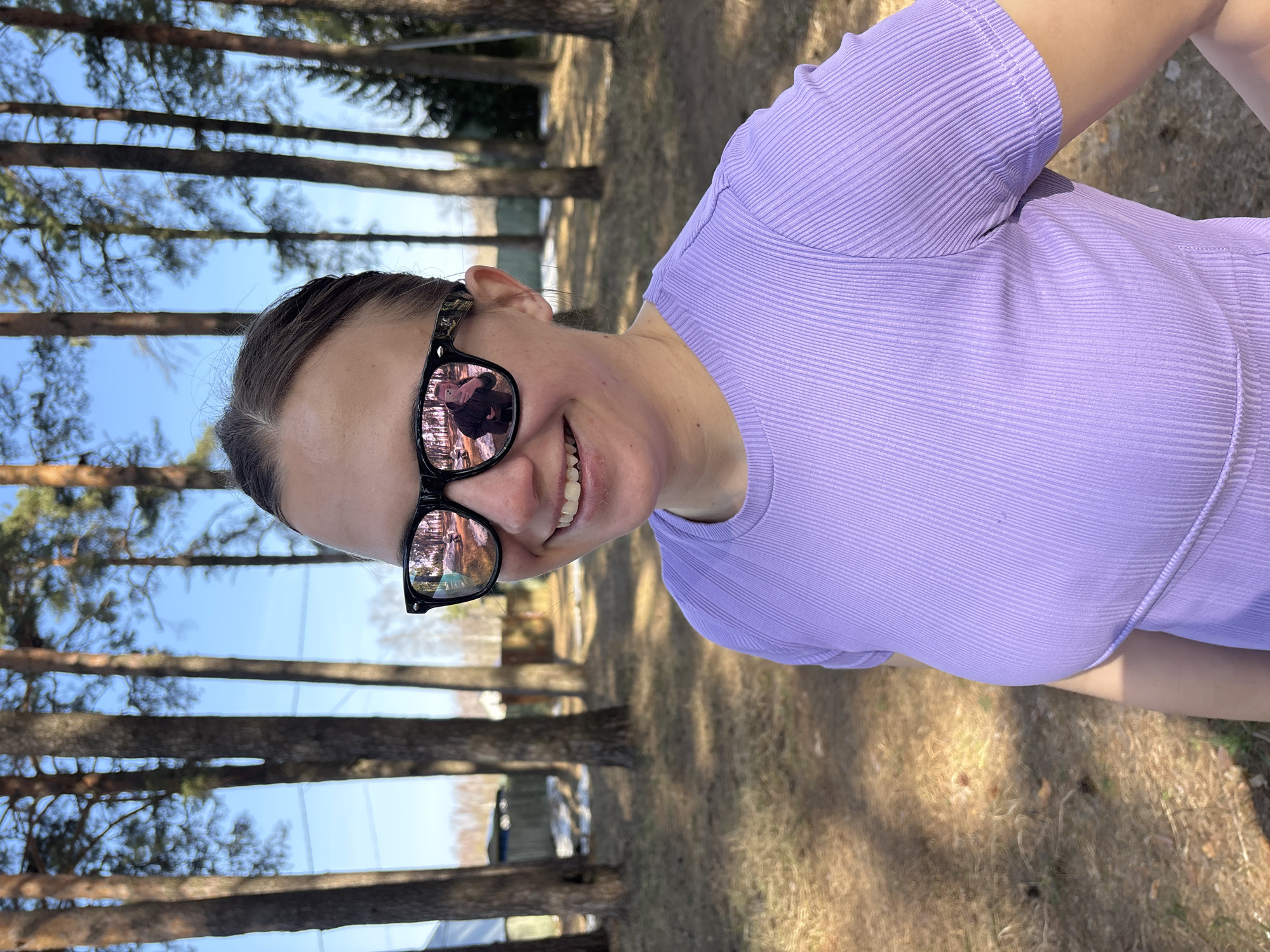 A female subject is depicted outdoors in the city of Раздолье, Russia, under clear, bright daylight. The subject, positioned centrally, is smiling with teeth visible and facing slightly to the left. She has dark hair pulled back from her face and wears black-framed sunglasses with reflective pink-orange lenses. Her attire consists of a light purple, ribbed, short-sleeved top. A reflection in her sunglasses shows a male figure, presumed to be the photographer, holding a mobile device and wearing a dark jacket. The background environment consists of numerous tall, slender coniferous trees with dark trunks. The ground is covered with dry, light-brown grasses and scattered patches of melting snow. In the distant left background, a light-colored building with a dark roof and a wooden fence is partially visible. The sky overhead is clear blue.