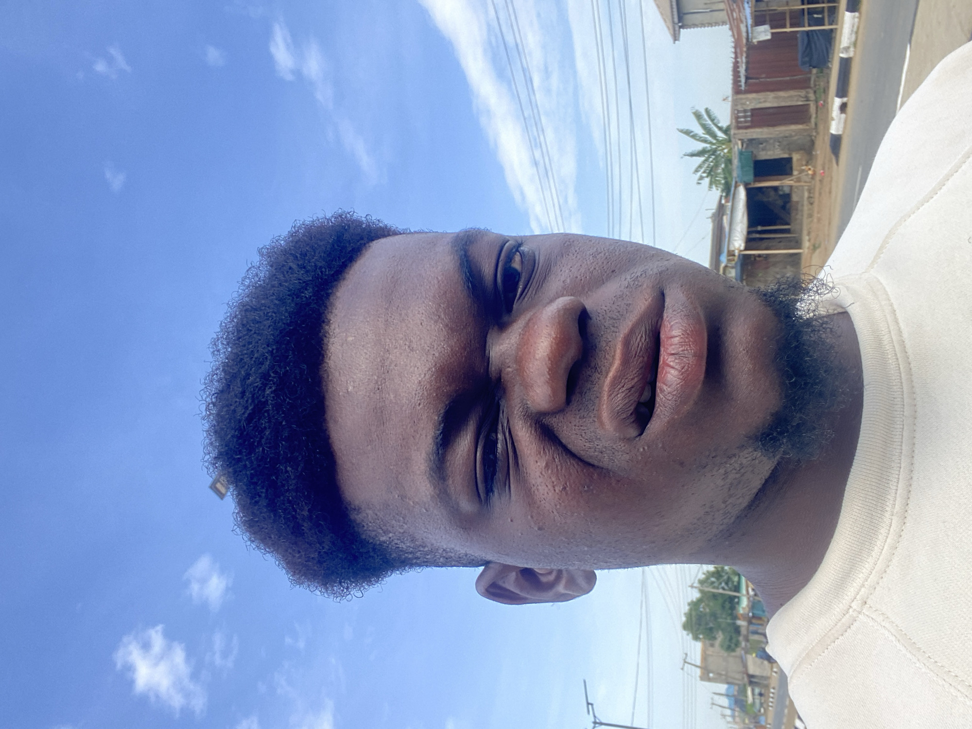 A close-up, slightly tilted selfie of a dark-skinned man with a short, curly afro and a trimmed beard. He wears a white crewneck shirt. The background includes a clear, blue sky with scattered clouds, telephone wires, and buildings along a street. The location appears to be an urban area in the city of Unknown, Unknown.