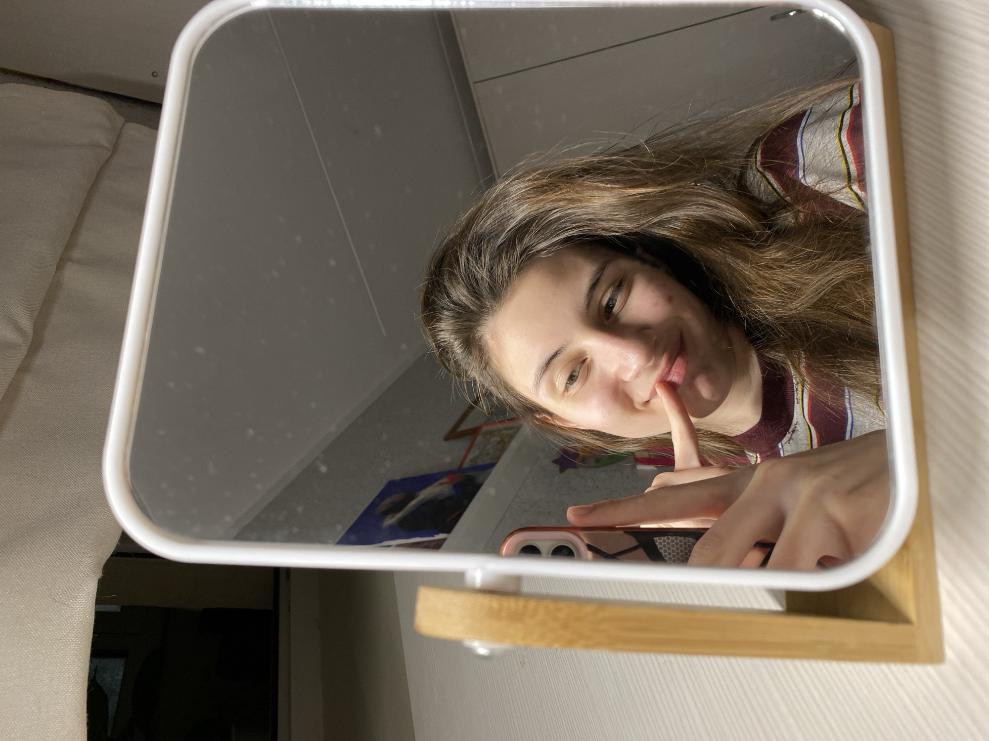 The image captures a young woman taking a mirror selfie in what appears to be a personal room in Votkinsk, Russia. She is reflected in a rectangular mirror with rounded white plastic edges, which rests on a light-colored wooden shelf. The woman has long, wavy brown hair and is wearing a striped shirt featuring red, blue, yellow, and white/grey bands. Her expression is a subtle, slightly playful smile, with her index finger gently touching her lower lip. Visible on her face are a few blemishes, notably on her forehead and left cheek.
She holds a rose gold or pink smartphone, partially visible in her hand, with its dual camera lenses reflecting in the mirror's lower left quadrant. The mirror surface itself shows numerous small dust specks and a faint diagonal scratch in the upper left corner.
The reflection in the mirror reveals parts of the room: a light-colored wall or ceiling in the upper sections, and to the left, a soft, light-colored fabric, likely a pillow or bedding. Below her face in the reflection, some indistinct objects are visible, including what appears to be a blue sheet or book with a dark image, and a star-shaped decoration further back. The physical background outside the mirror shows a light-colored, subtly patterned wallpaper or wall covering to the right and a similar light-colored fabric to the left. The lighting is soft and even, suggesting an indoor setting, possibly during the day or with artificial illumination. There is no discernible text visible in the image. The overall mood is casual and intimate, capturing a moment of self-reflection or a shared personal snapshot.