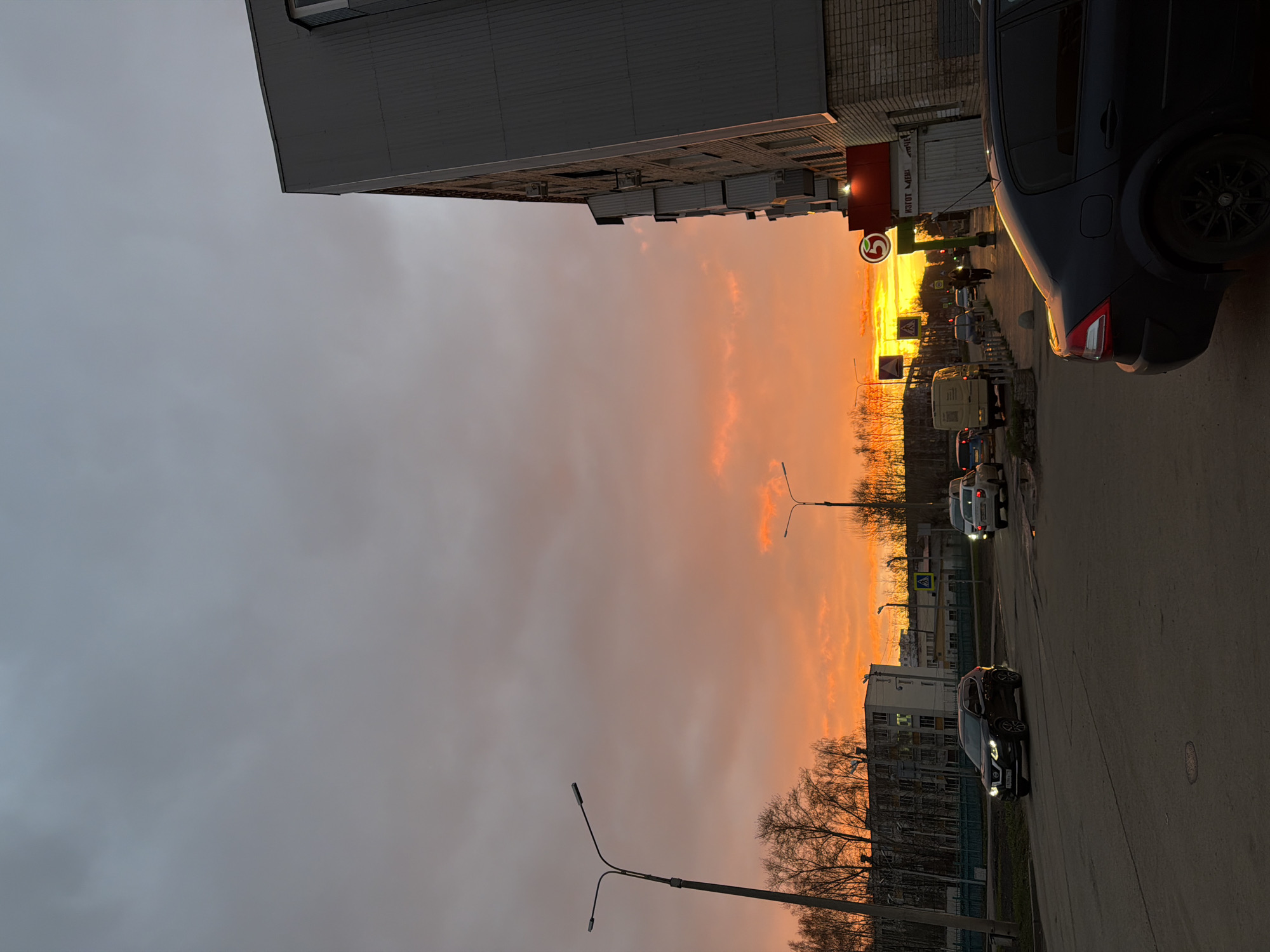 This media file captures an urban street scene at either sunrise or sunset, characterized by a dramatically colored sky transitioning from grey overcast on the left to vibrant orange and yellow near the horizon on the right. The image is composed with the horizon cutting diagonally across, with the sky dominating the upper-left and the street, buildings, and vehicles occupying the lower-right.
In the foreground, a dark-colored SUV or car is partially visible on the right side of the road. Further down the street, several other vehicles are present, including a white van and a black SUV with its headlights on, driving towards the viewer. A few indistinct figures appear to be walking or crossing the street in the distance.
On the right side of the road, there's a building with a prominent red and white sign featuring a large "5" logo, indicative of the Russian Pyaterochka supermarket chain. Adjacent to or on this structure, additional text reads "ИЗГОТОВЛЕНИЕ МЕБЕЛИ" (Manufacturing Furniture). Traffic signs, including a pedestrian crossing sign, are also visible. Bare trees, possibly indicating autumn or winter, silhouette against the bright sky. The license plate on the black SUV has the characters "K208YY" and region code "116", which corresponds to the Republic of Tatarstan, confirming the location in Naberezhnye Chelny, Russia.
The time of day is clearly dawn or dusk, with low, warm light casting long shadows and creating a strong contrast with the dimmer, grey clouds overhead. The weather appears overcast but with a break near the horizon allowing the sun's colors to be seen. The scene depicts everyday activity on a city street under a striking sky.