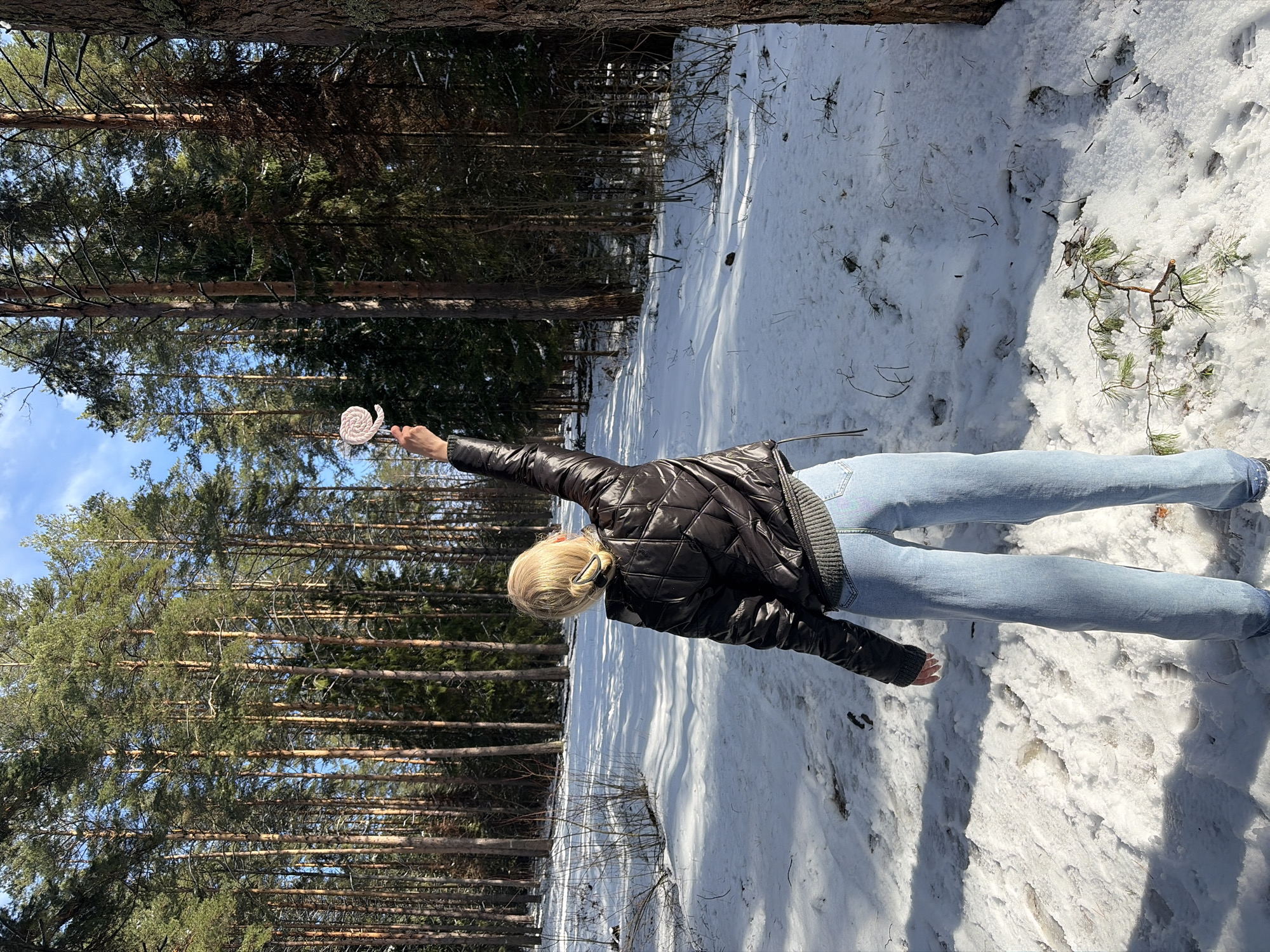 The image shows a woman lying on her back in the snow in a forest in Прикамский, Russia. She is wearing jeans and a black quilted jacket, and has blonde hair pulled back. Her right arm is extended, holding a white swirl lollipop up towards the sky. The background features tall pine trees and a snowy ground. The lighting suggests it is a sunny day.