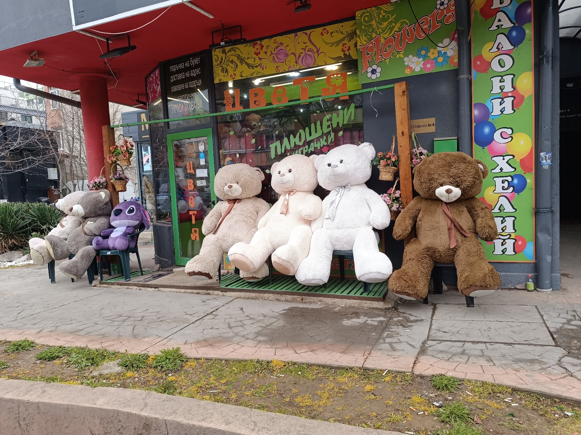 Here is a detailed description of the image:
**Scene:** A storefront of a flower and plush toy shop.
**Location:** Sofia, Bulgaria (indicated by Cyrillic text on storefronts).
**Subjects/Objects:**
* Five stuffed toys (four teddy bears of varying colors and sizes, and one purple character) seated on chairs outside the storefront.
* Flower shop storefront with "Flowers" and "Plush Toys" signs in Cyrillic.
* A sign with "Balloons" in Cyrillic with the image of multicolored balloons.
* A table with flowers in pots.
* Green grate bench.
* Concrete sidewalk and curb with weeds growing.
* Red awning and building facade.
**Environment:**
* Urban street scene with architectural elements.
* Gray sky visible.
* Bare tree branches are visible in the background, indicating either late autumn, winter, or early spring.