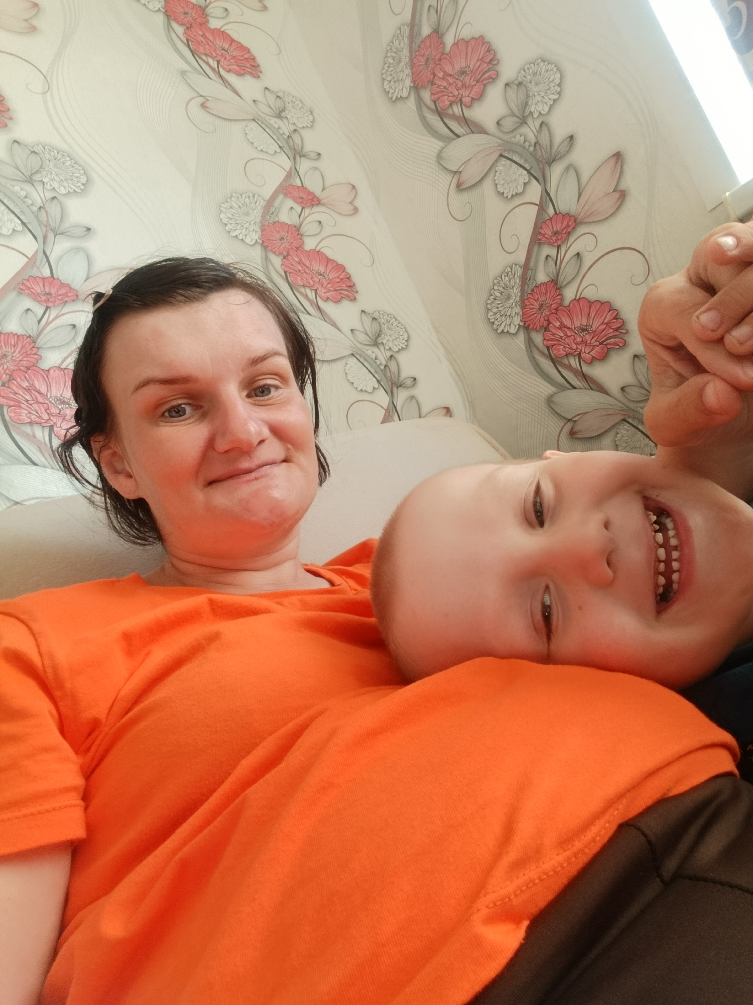 A selfie captured at Фоки, Russia, shows a woman and child posing against a floral wallpaper backdrop. The woman, on the left, has dark hair and a slight smile. She is wearing an orange T-shirt. The child, with a shaved head, is lying on her chest and smiling broadly, showcasing a set of baby teeth. A partial view of a hand is visible in the upper right corner.