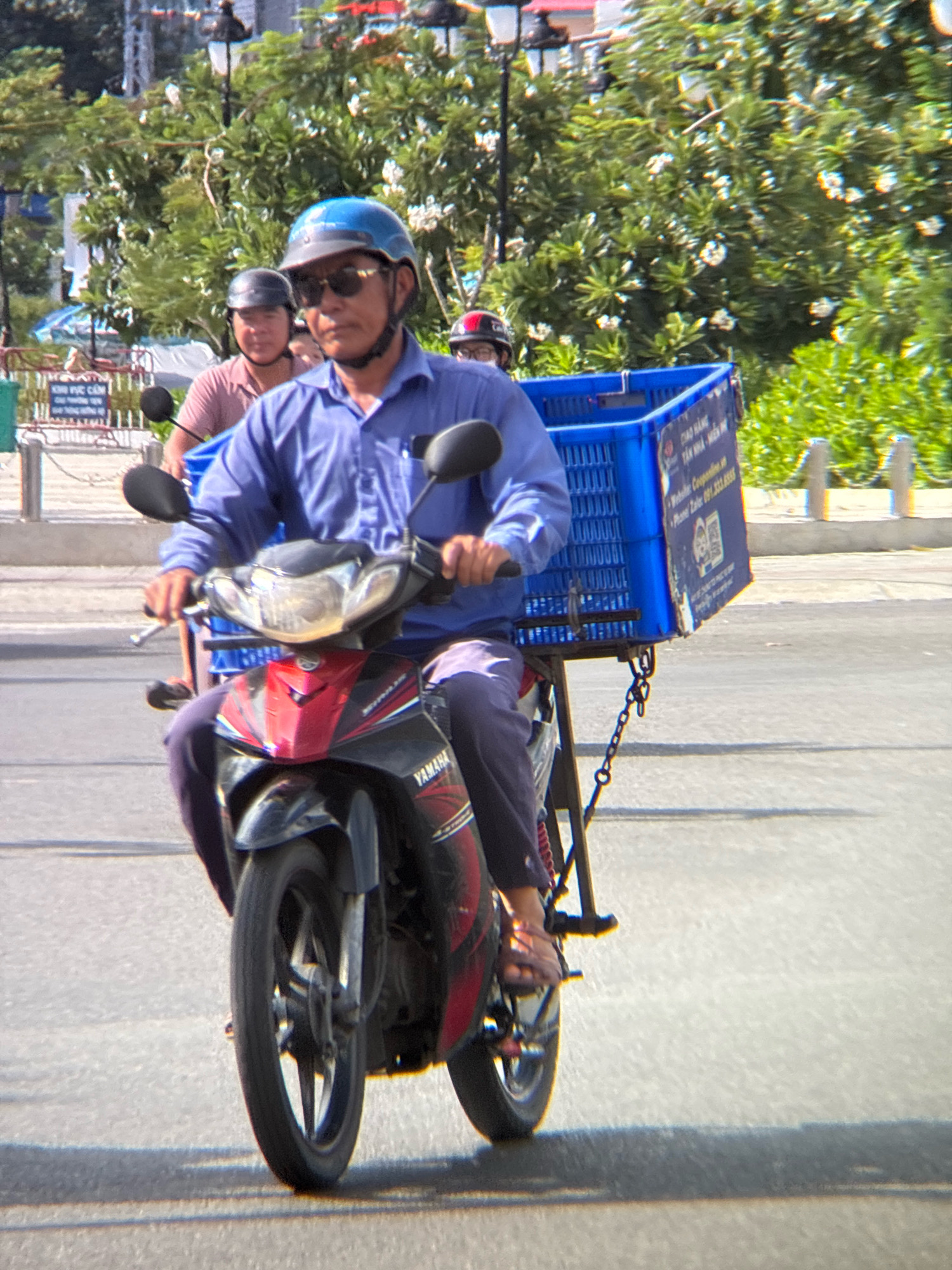 A man wearing a blue helmet and sunglasses is riding a red and black Yamaha motorcycle on a paved road in Nha Trang, Vietnam. He is wearing a blue collared shirt and dark pants. A large blue plastic basket is attached to the back of the motorcycle, secured with a chain. The basket has some writing on it, including what appears to be a website and phone number. In the background, another person on a motorcycle is visible, wearing a dark helmet and a pink shirt. Trees with white flowers are in the background, along with some streetlights and a fence. The image is taken from a slightly low angle, capturing the motorcycle and its rider in motion.