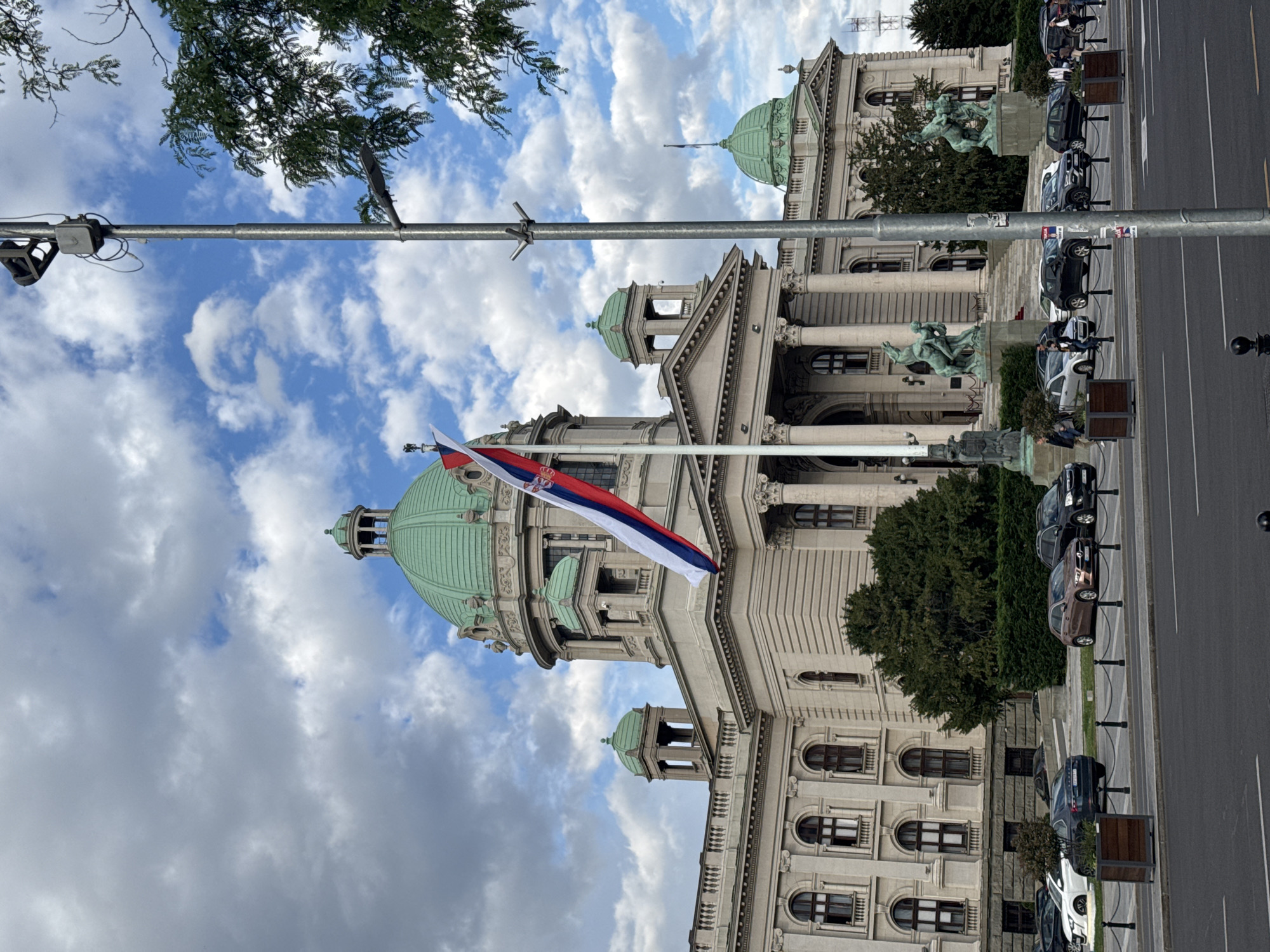 The image captures the National Assembly of Serbia in Belgrade, Serbia, under a partly cloudy sky. The Serbian flag is prominently displayed in front of the building. The architectural details of the parliament building are visible, including its domes, columns, and windows. A street with parked cars runs alongside the building. A lamppost is visible in the foreground. There are two statues of men on horses in front of the building. There are people on foot next to the parked cars.
